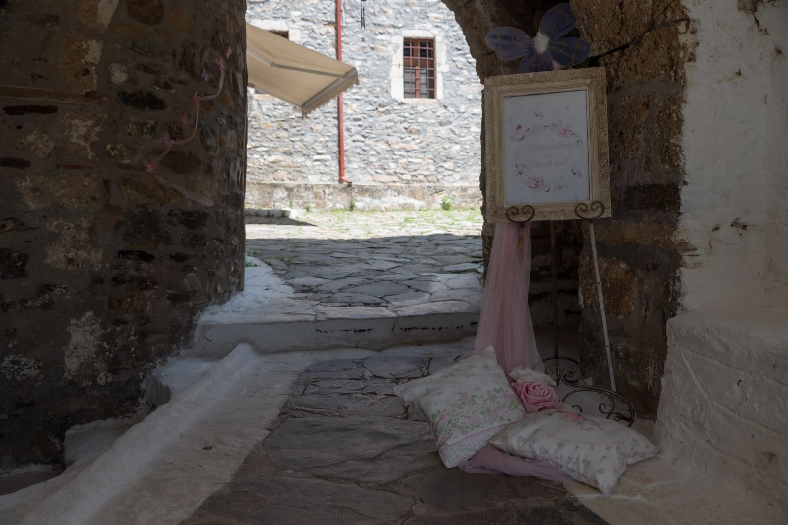 A stone passage with a framed sign on an easel, pink fabric and cushions on the paving.