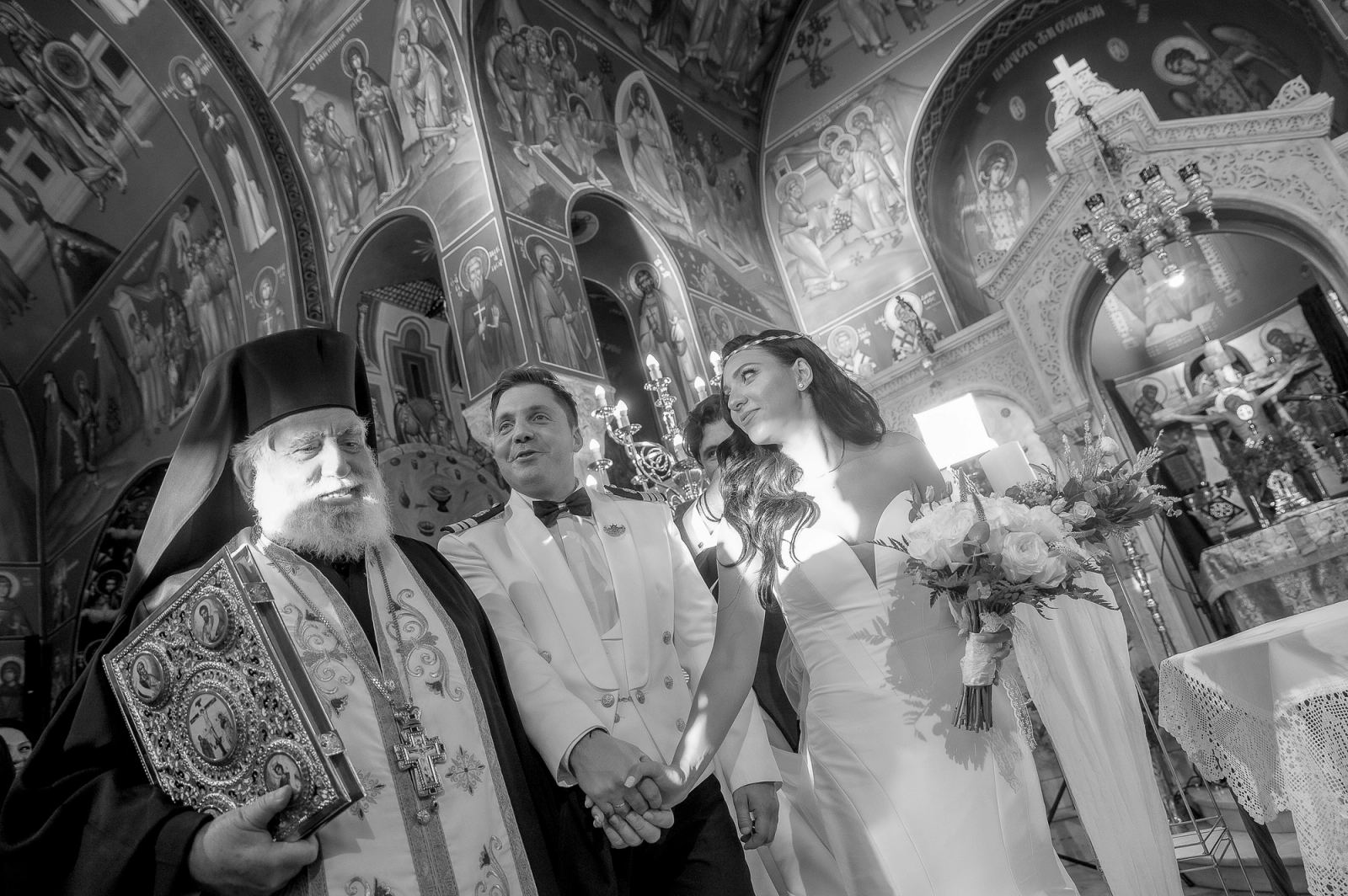 Ασπρόμαυρη λήψη του ιερέα με Ευαγγέλιο δίπλα στη νύφη και τον γαμπρό με λευκή στολή, πιασμένους χέρι χέρι.