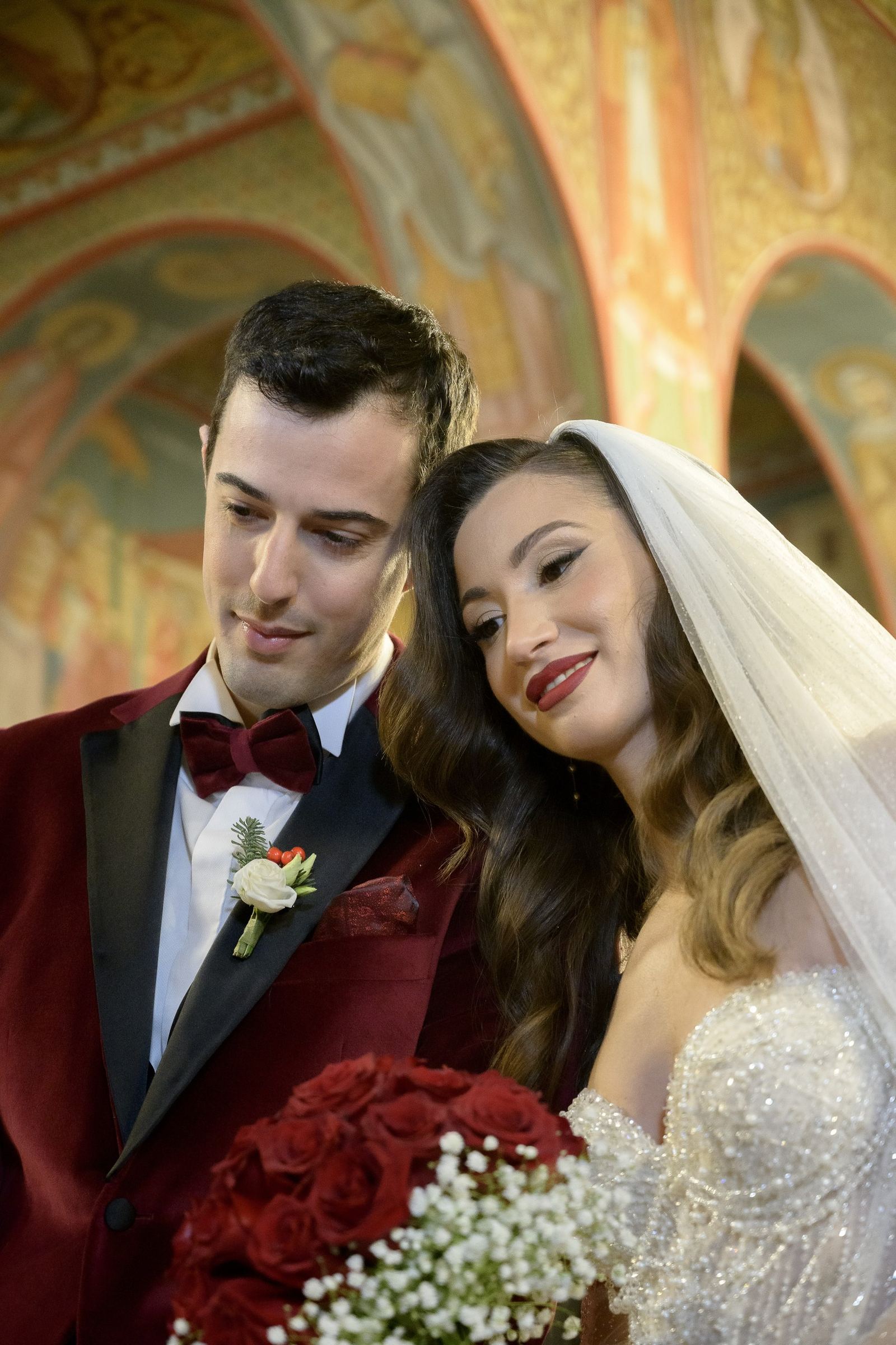 The bride leans on the groom holding red roses beneath church frescoes