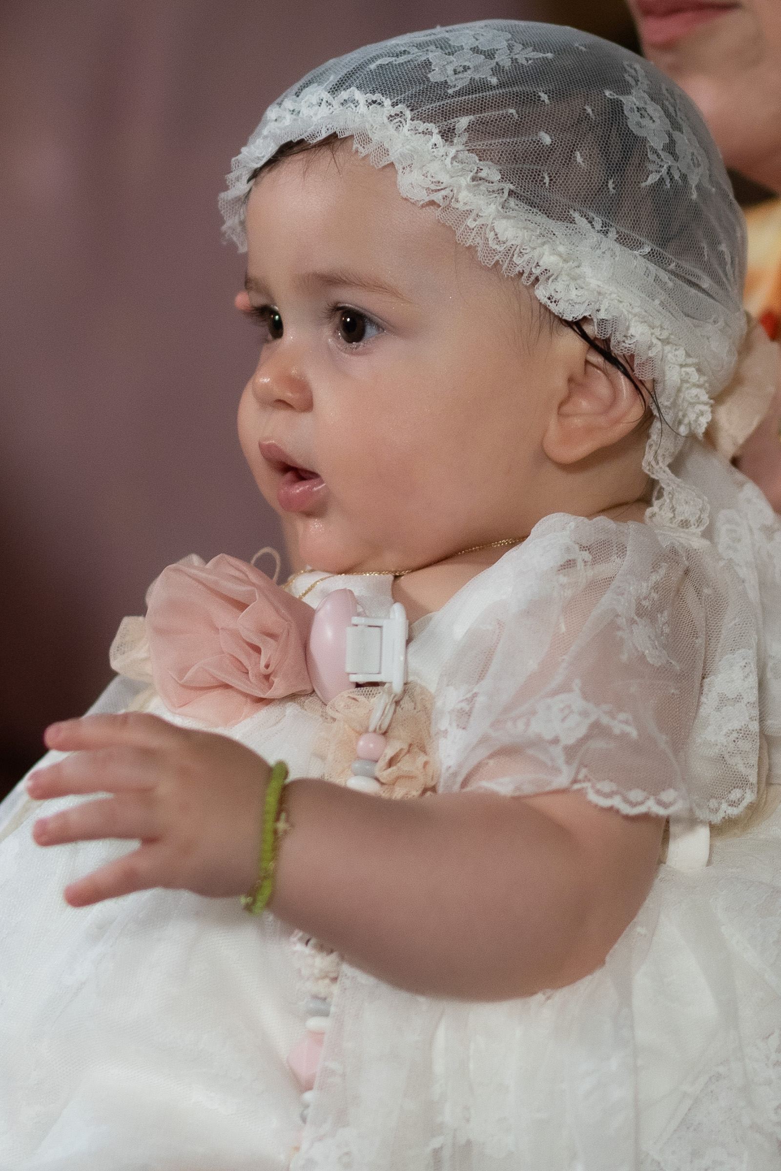 The baby in a white lace dress and bonnet looks to the side.