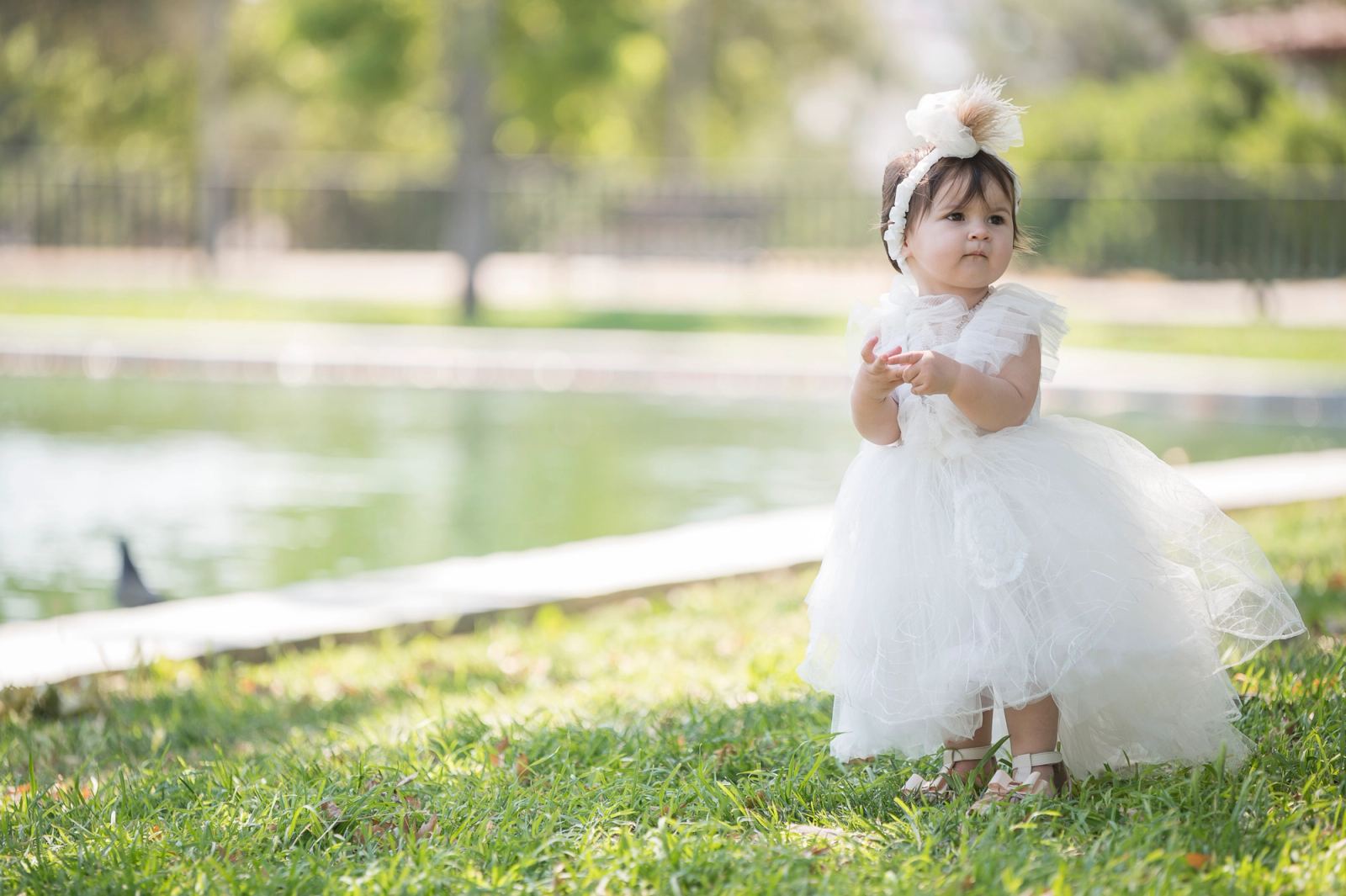 The baby in a white dress and hair bow stands on the grass in front of a pond.