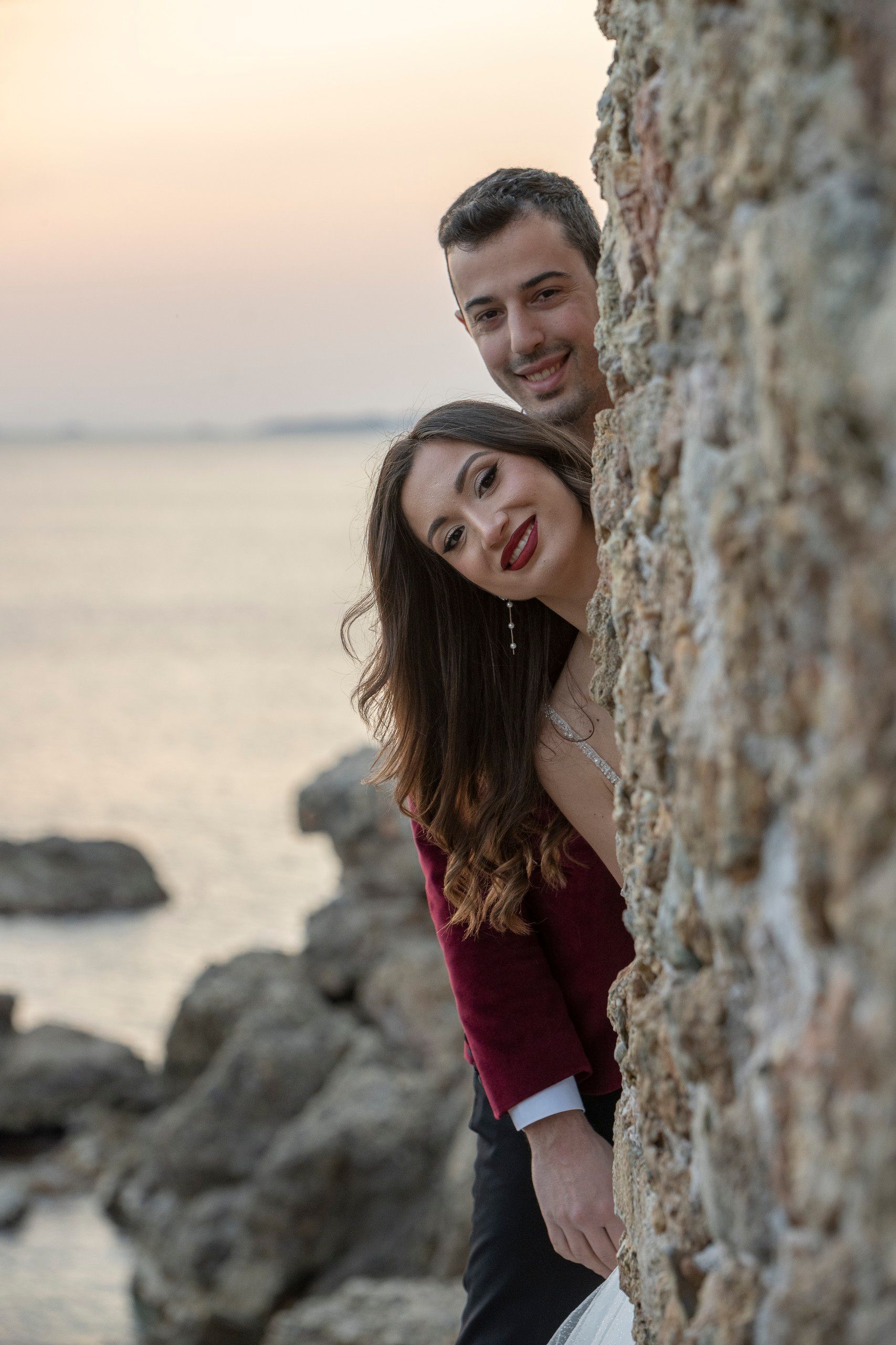 Bride and groom peeking out from behind a stone wall by the sea