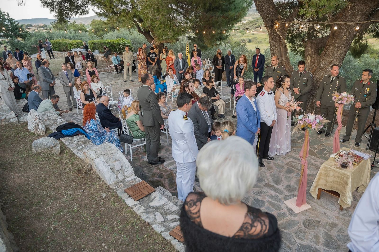 Υπαίθριος γάμος σε πέτρινη αυλή με ελιές, καλεσμένους γύρω και στρατιωτικούς δίπλα στο ζευγάρι.
