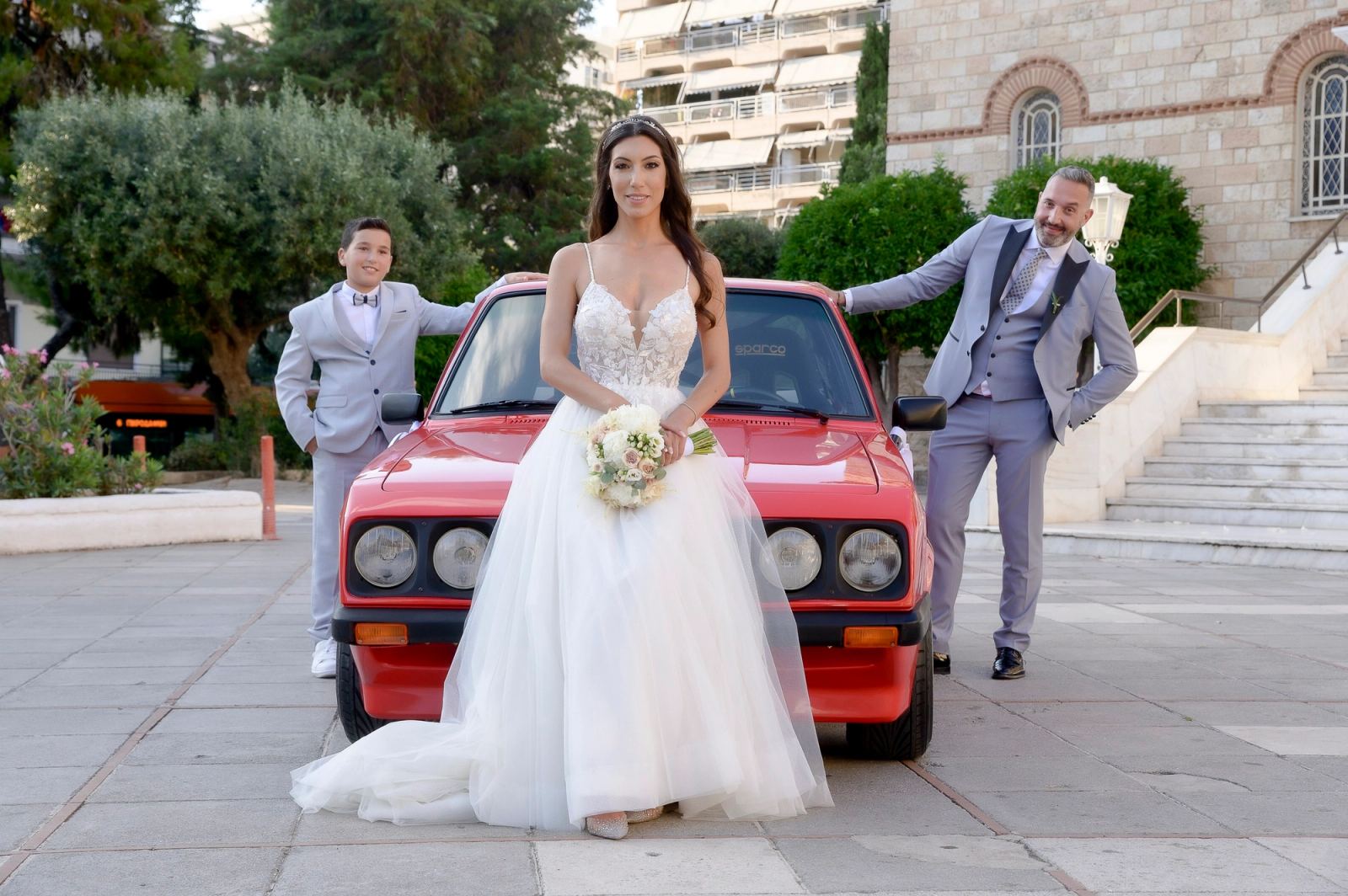 Η νύφη με ανθοδέσμη μπροστά σε κόκκινο κλασικό αυτοκίνητο, με τον γαμπρό και ένα αγόρι με γκρι κοστούμια δίπλα.