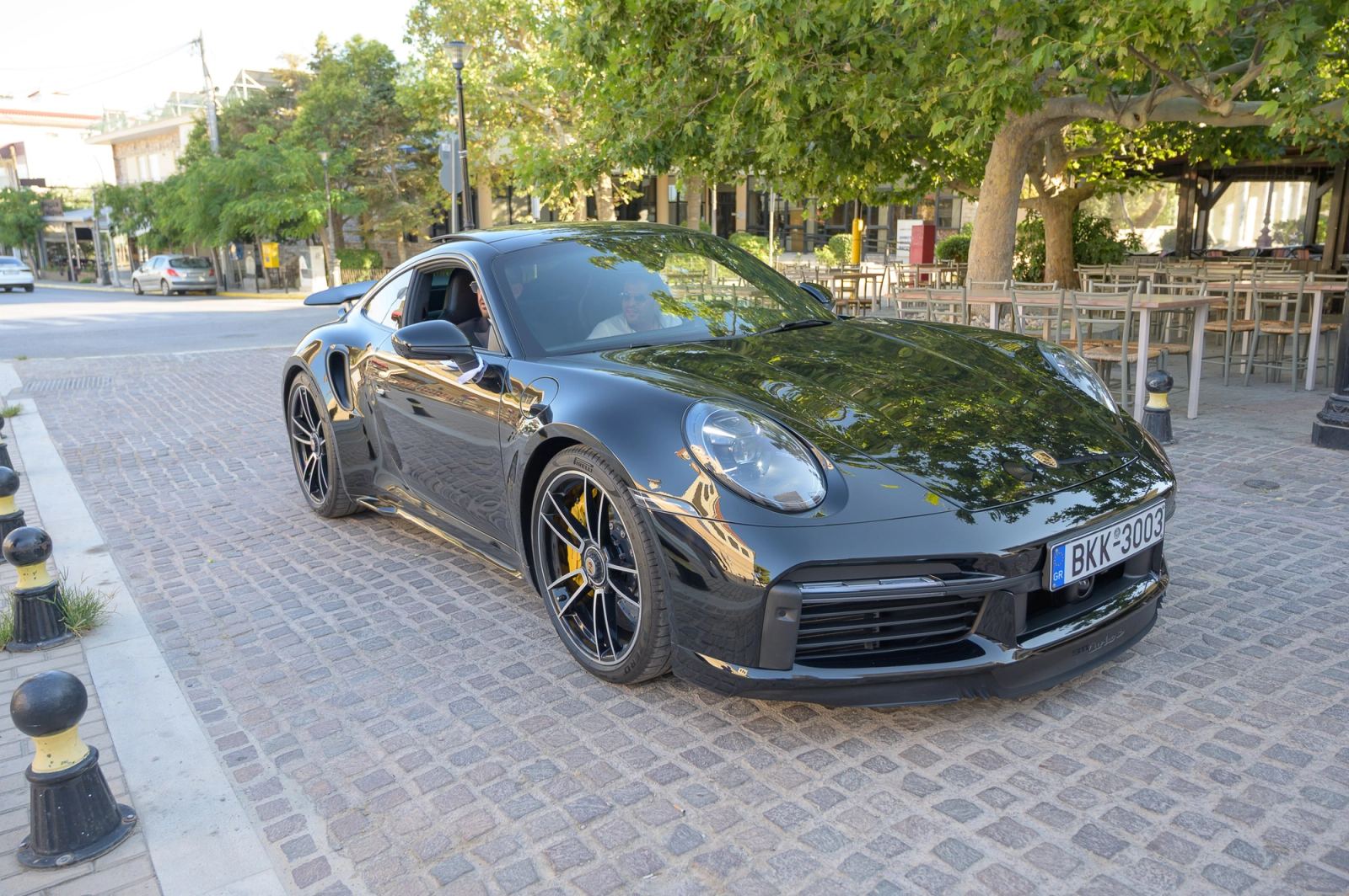 A black sports car on a cobbled street under trees, a driver in sunglasses at the wheel.