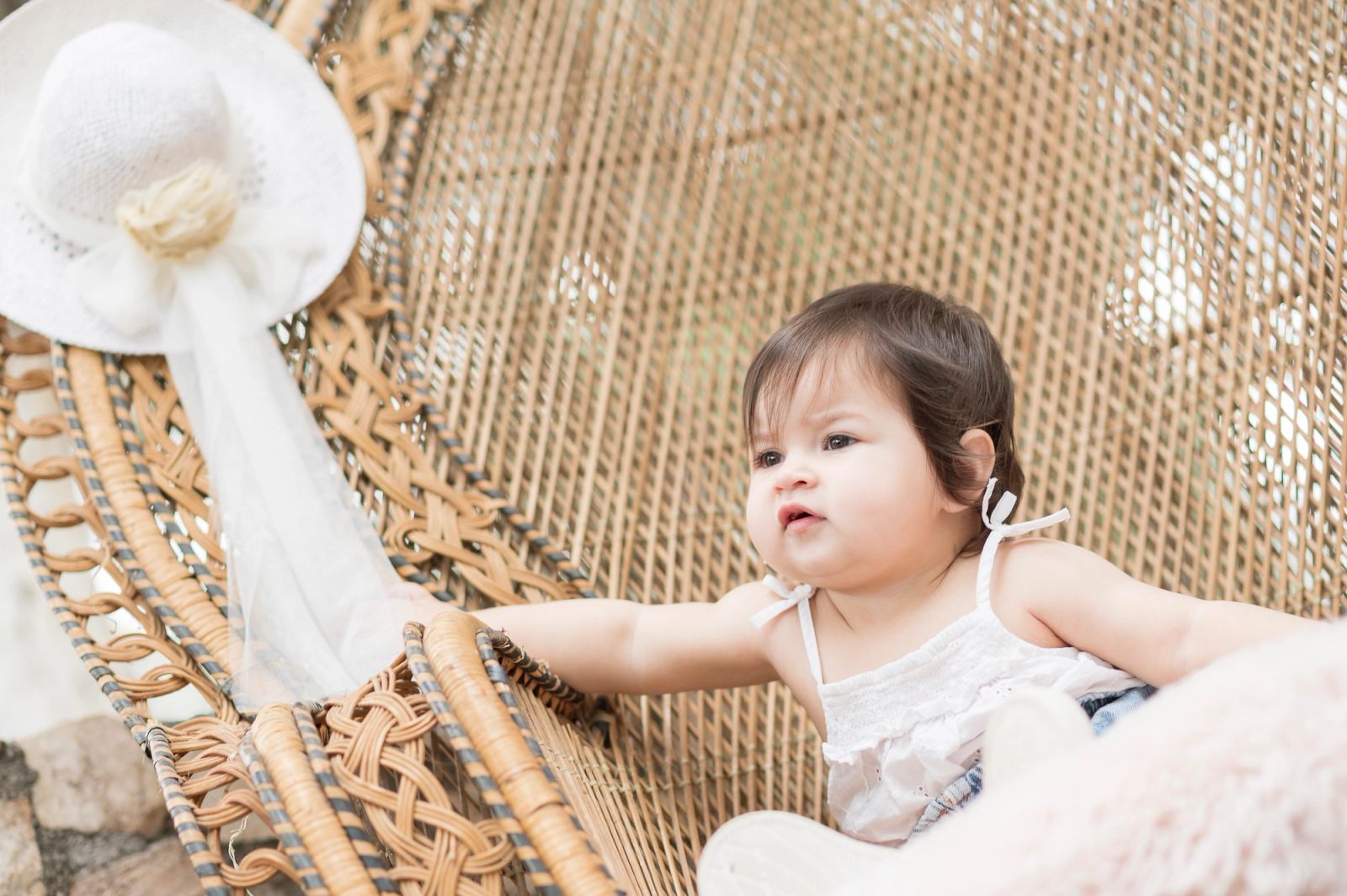 The baby sits in a wicker peacock chair with a white hat hanging on its edge.