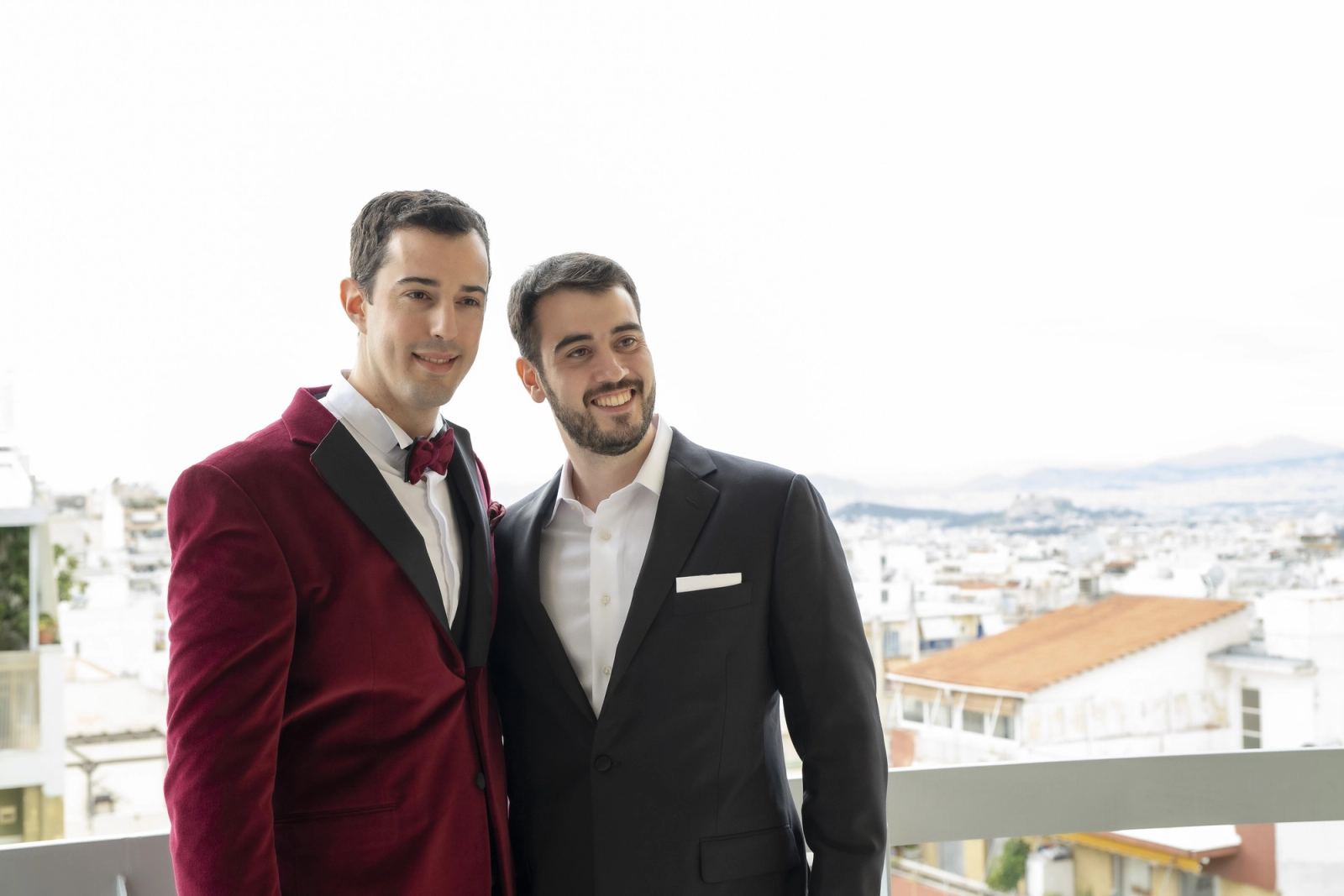 The groom in a burgundy velvet jacket and a man in a black suit on a balcony overlooking the city