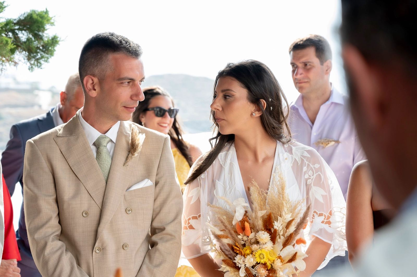 Ο γαμπρός και η νύφη κοιτάζονται στην υπαίθρια τελετή, με καλεσμένους πίσω τους.