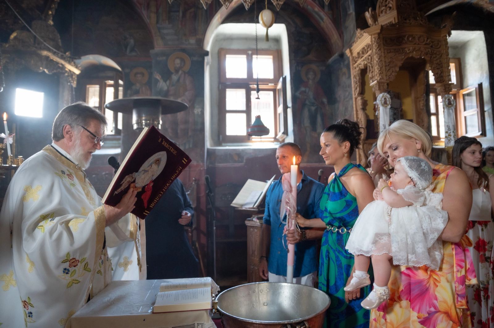 The priest reads from a red book before guests with a candle and a woman holding the baby in white.