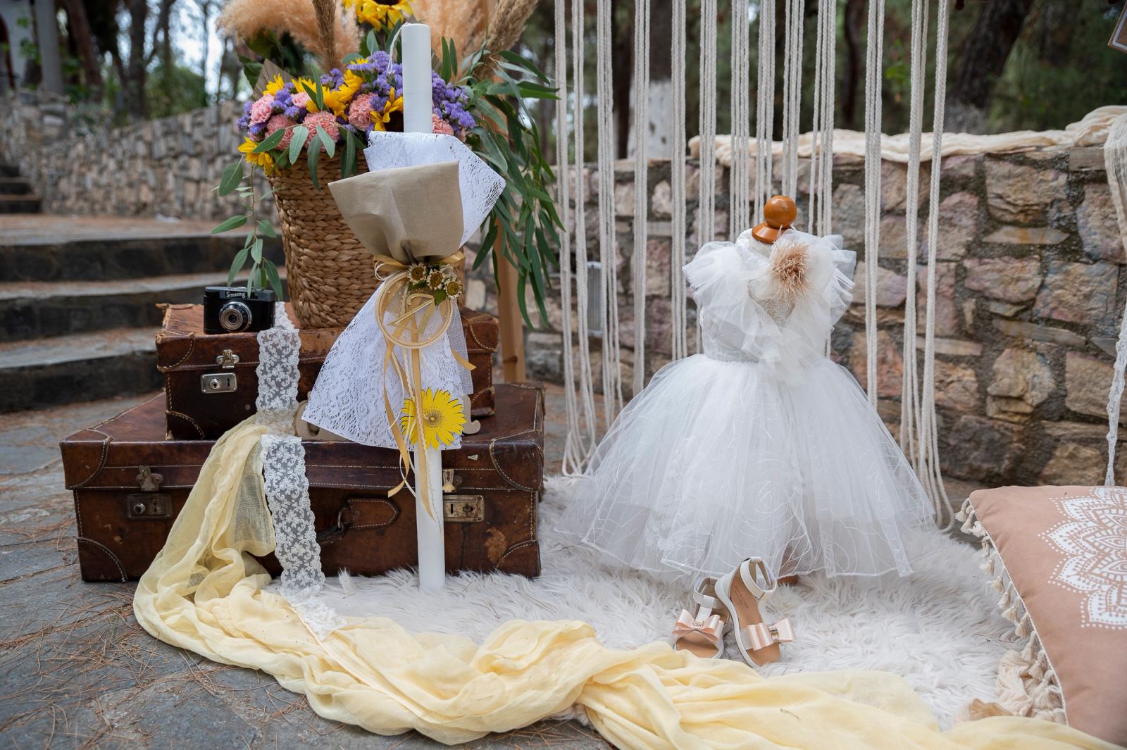 White christening dress on a stand, a candle, vintage suitcases and a flower basket before a stone wall.