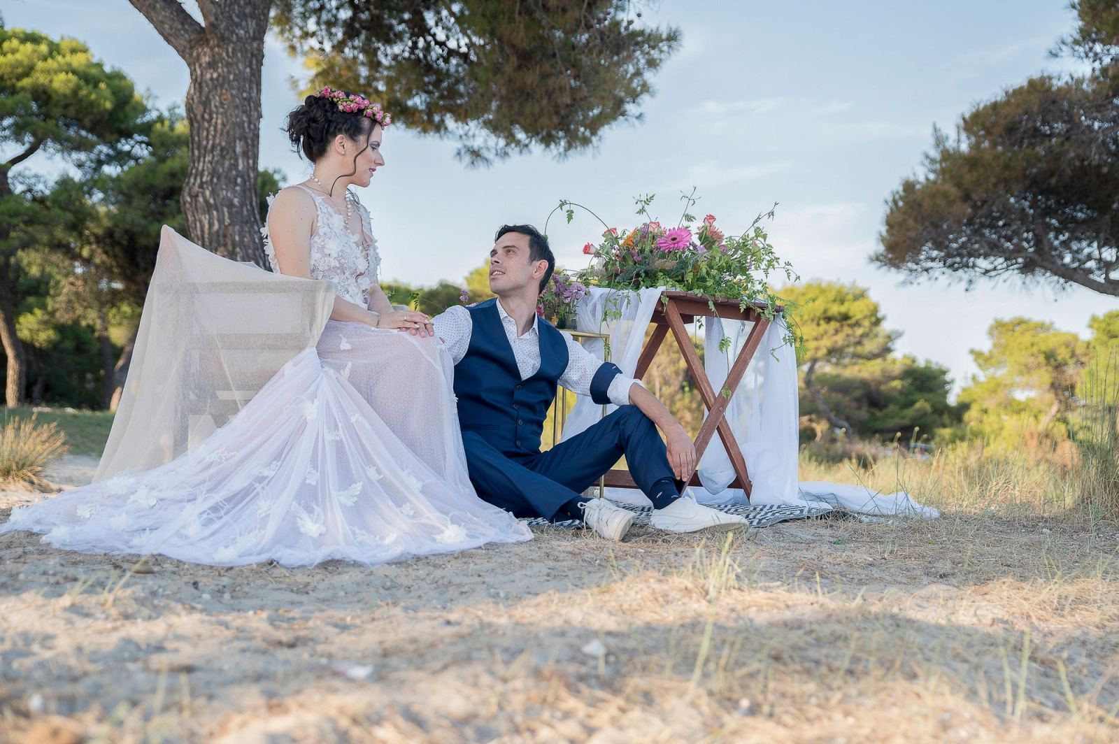 Η νύφη με στεφάνι από λουλούδια κάθεται και ο γαμπρός στο χώμα δίπλα της, κάτω από πεύκα, με τραπεζάκι με λουλούδια.