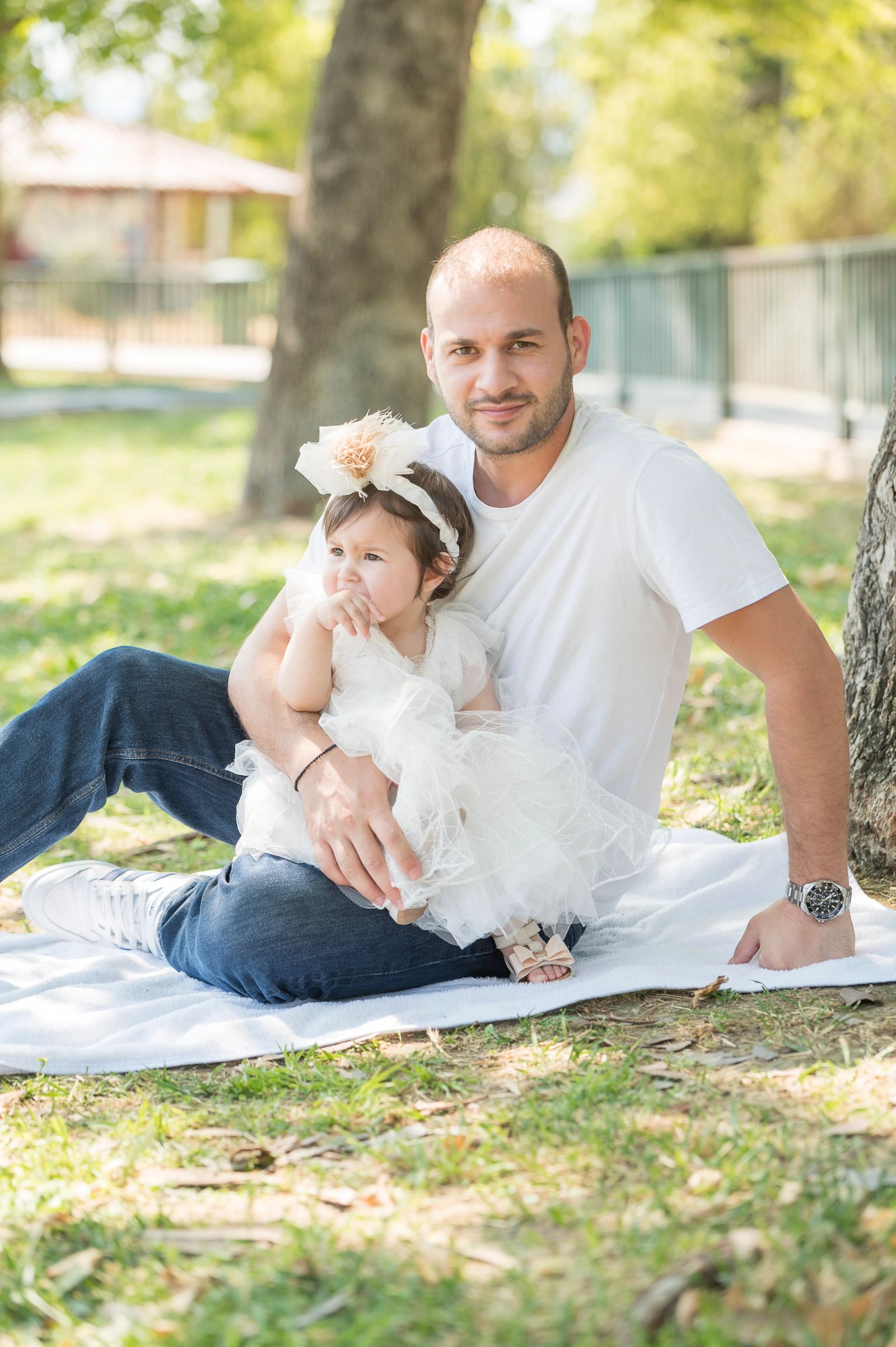 A man in a white T-shirt sits on a blanket on the grass holding the baby in a white dress.