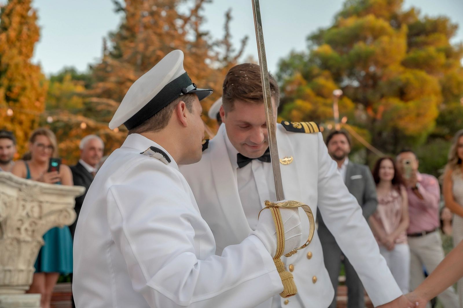 Αξιωματικός με πηλίκιο και λευκά γάντια σηκώνει σπαθί πάνω από τον γαμπρό, ενώ καλεσμένοι τραβούν με κινητά.