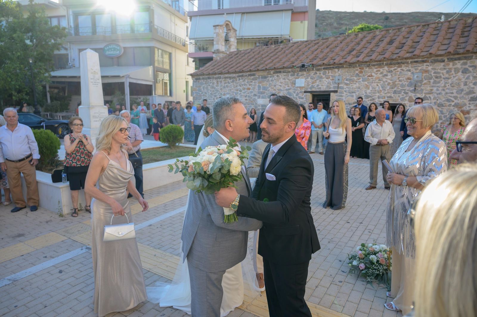 The groom embraces a man in a grey suit holding a bouquet outside a stone church, guests around them.