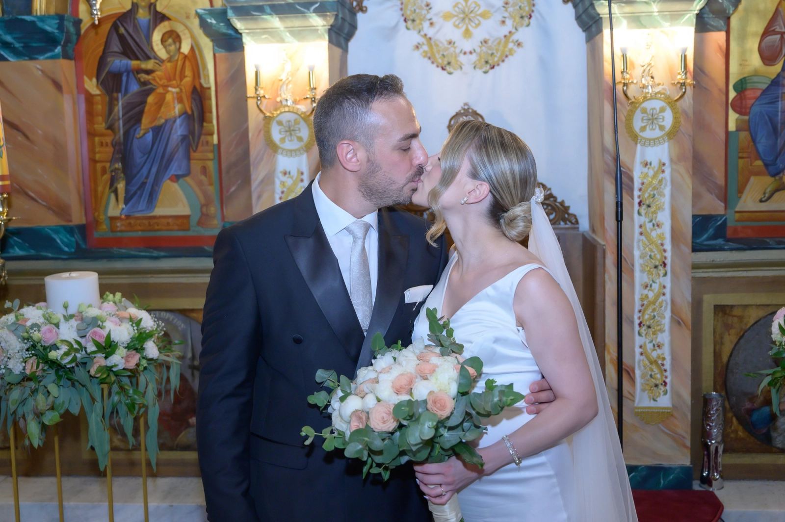 The bride and groom kiss before the iconostasis, she holding a bouquet of white and peach roses.