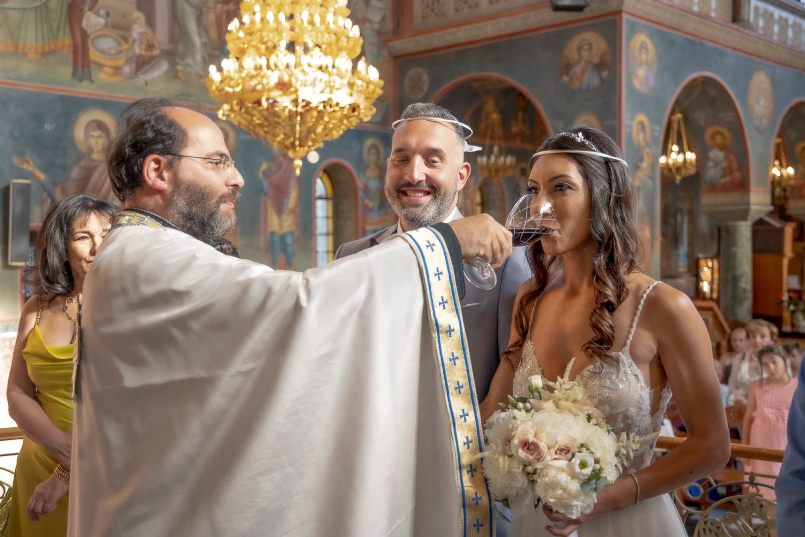 Η νύφη πίνει κρασί από ποτήρι που κρατά ο ιερέας, ενώ ο γαμπρός χαμογελά κάτω από χρυσό πολυέλαιο.