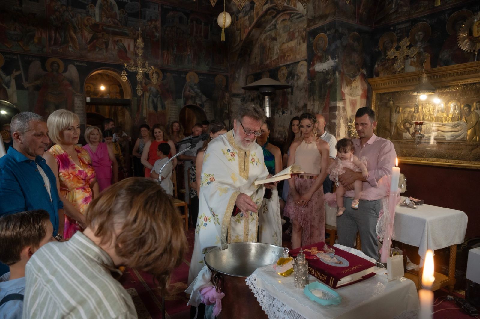 The priest reads beside the font in a frescoed church, with guests and a man holding the baby.