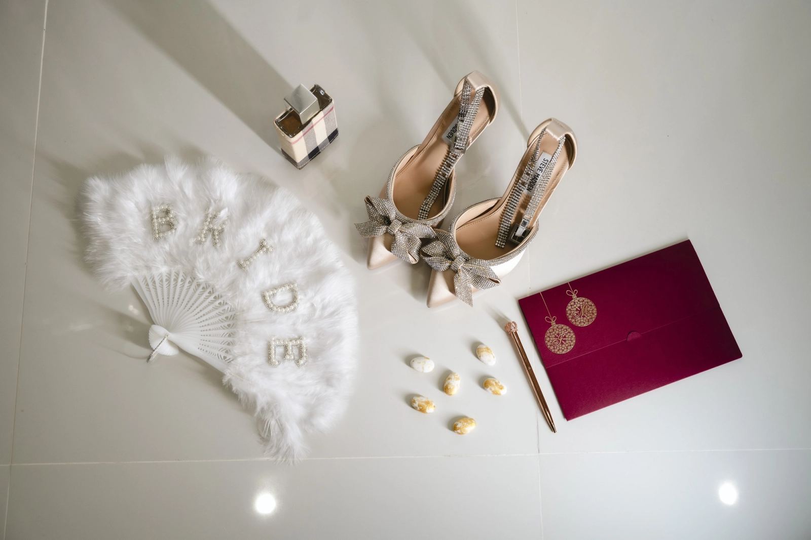 Bridal heels with rhinestone bows, a feather fan spelling BRIDE, perfume and a burgundy invitation on a white floor