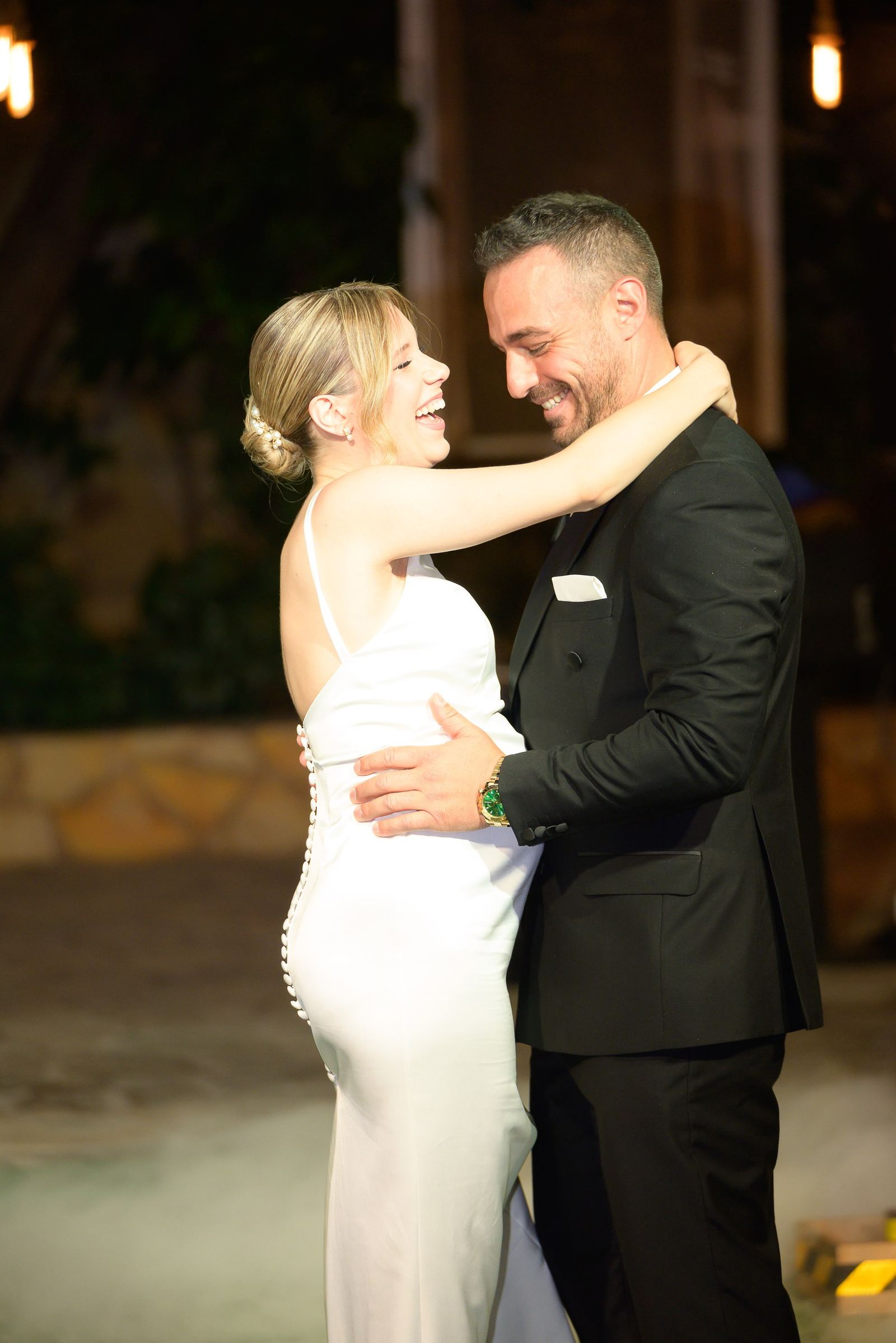 The bride laughs with her arms around the groom's neck as they dance at night.