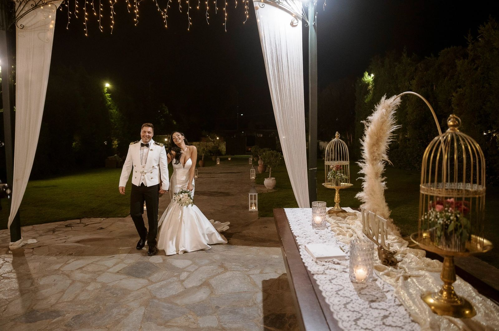 Ο γαμπρός και η νύφη περπατούν τη νύχτα κάτω από φωτάκια, δίπλα σε τραπέζι με χρυσά κλουβιά και κεριά.