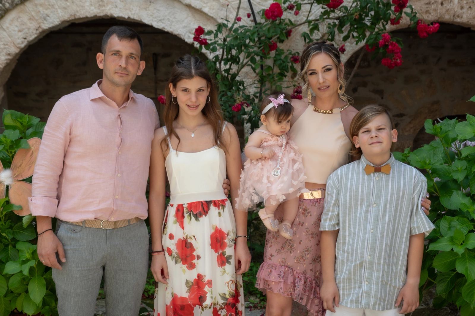 A family holding the baby in a pink tulle dress poses before stone arches with red roses and green plants.