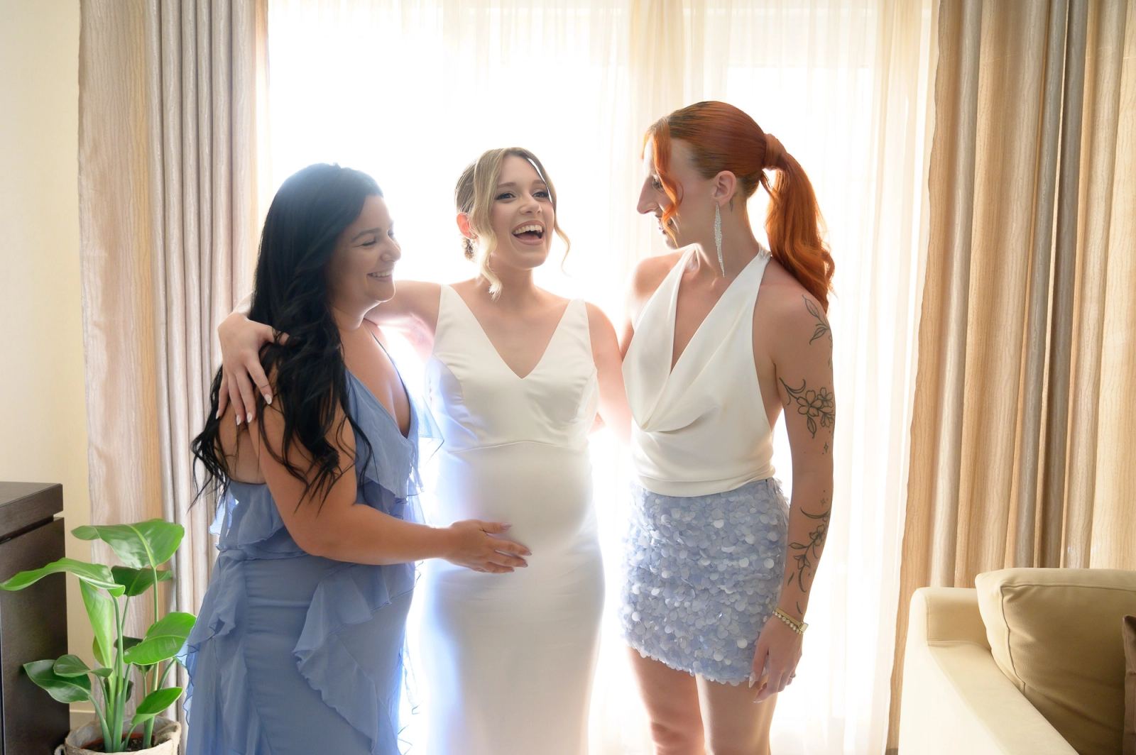 The bride laughs, arms around two friends in blue and white, in front of a sunlit window.