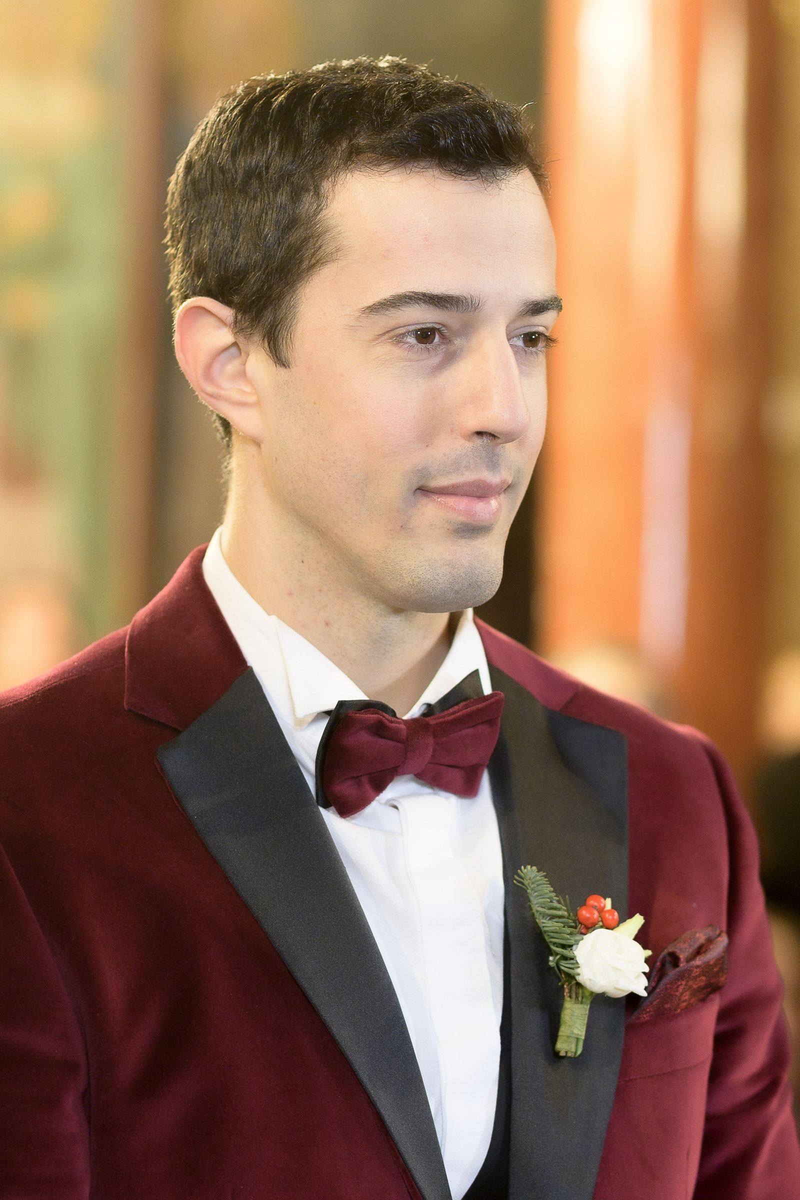 The groom in a burgundy velvet tuxedo, bow tie and boutonniere inside a church