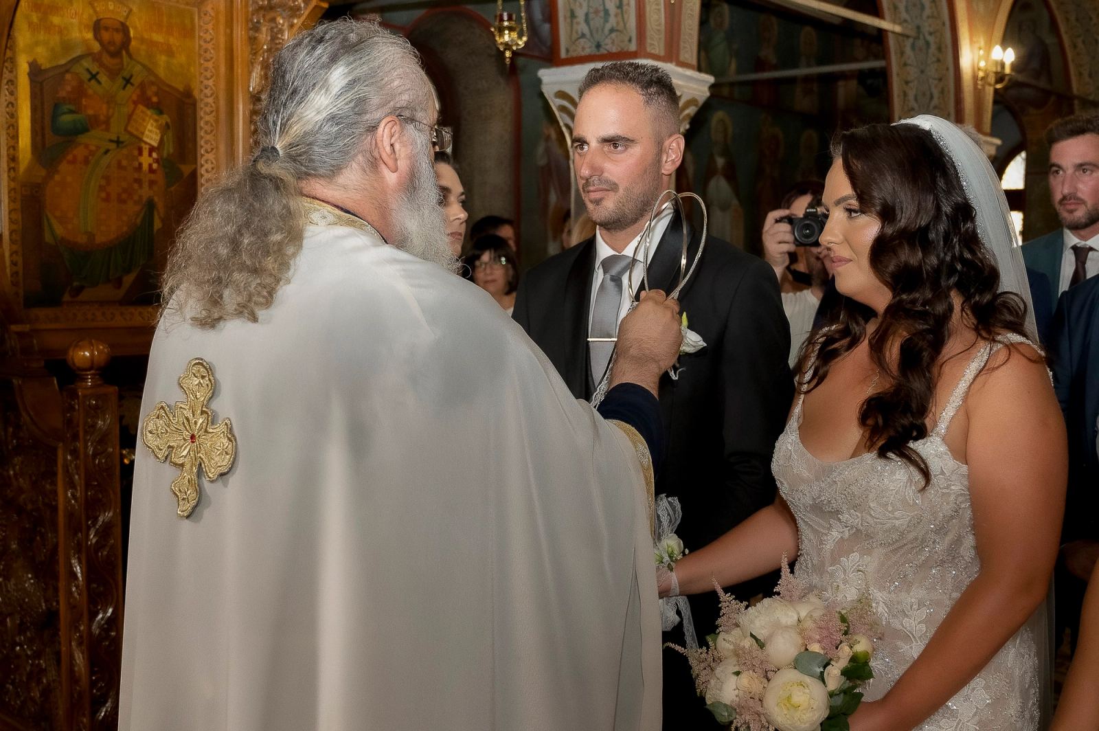 Ο ιερέας με λευκά άμφια κρατά τα στέφανα μπροστά στη νύφη και τον γαμπρό μέσα στην εκκλησία.