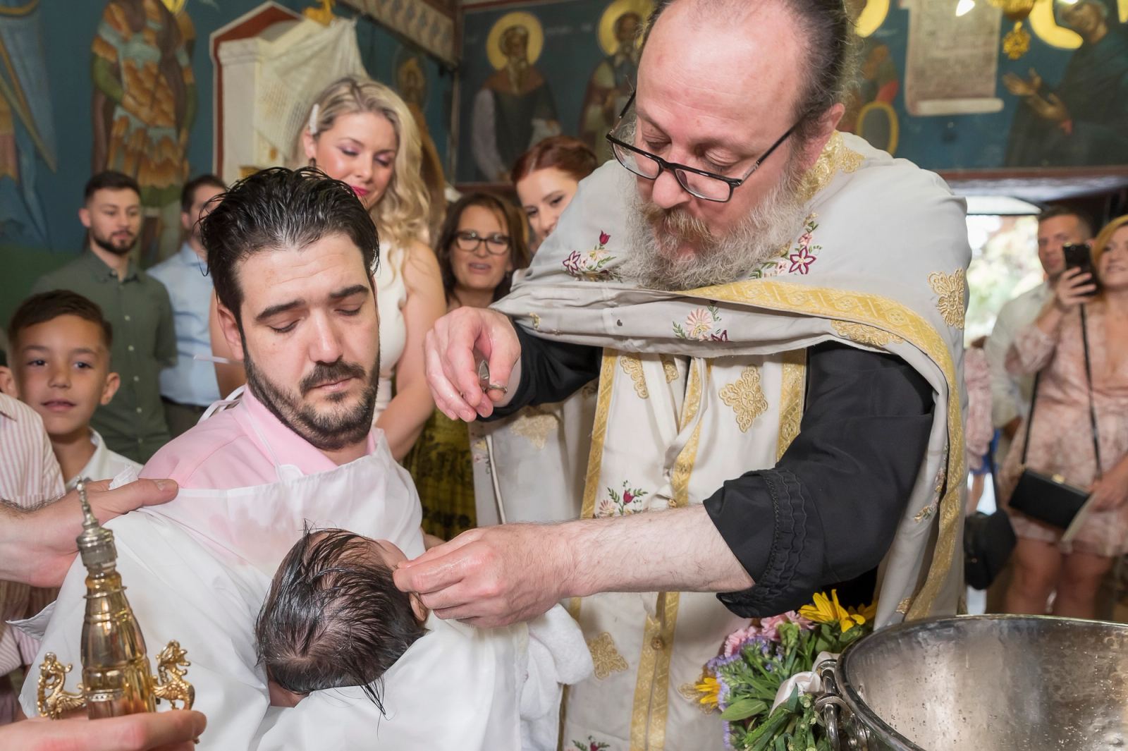 The priest anoints the baby held by a man in a white apron, with guests behind them.