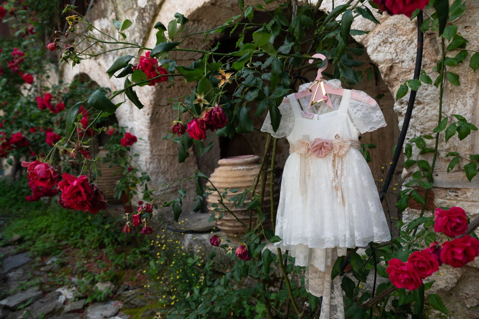 A white christening dress hangs among red roses in front of a stone arch.