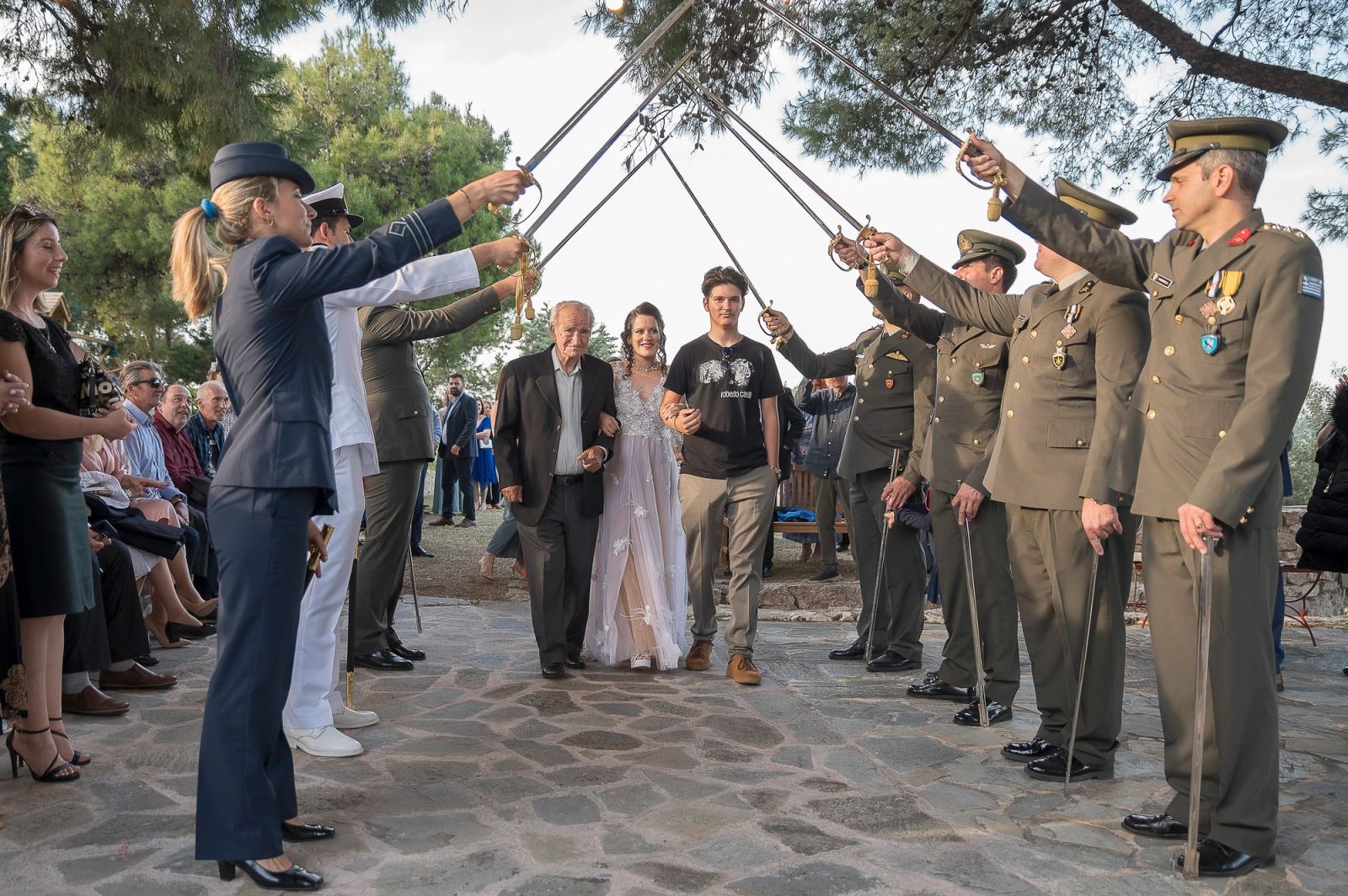 Η νύφη περπατά ανάμεσα σε δύο άνδρες κάτω από αψίδα από σπαθιά στρατιωτικών με στολή σε πέτρινη αυλή.