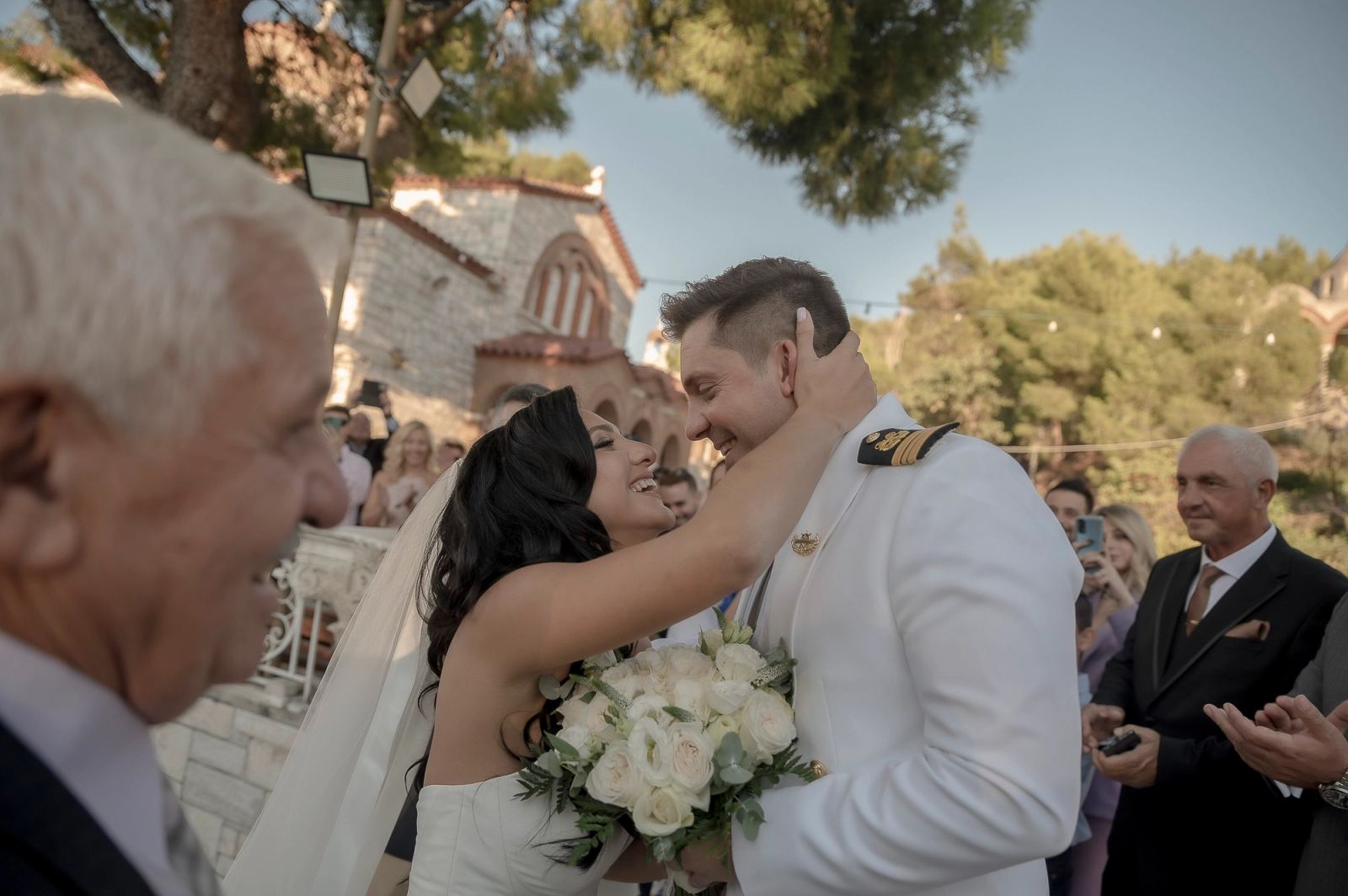 Η νύφη με ανθοδέσμη αγκαλιάζει τον γαμπρό έξω από πέτρινη εκκλησία, με καλεσμένους γύρω.