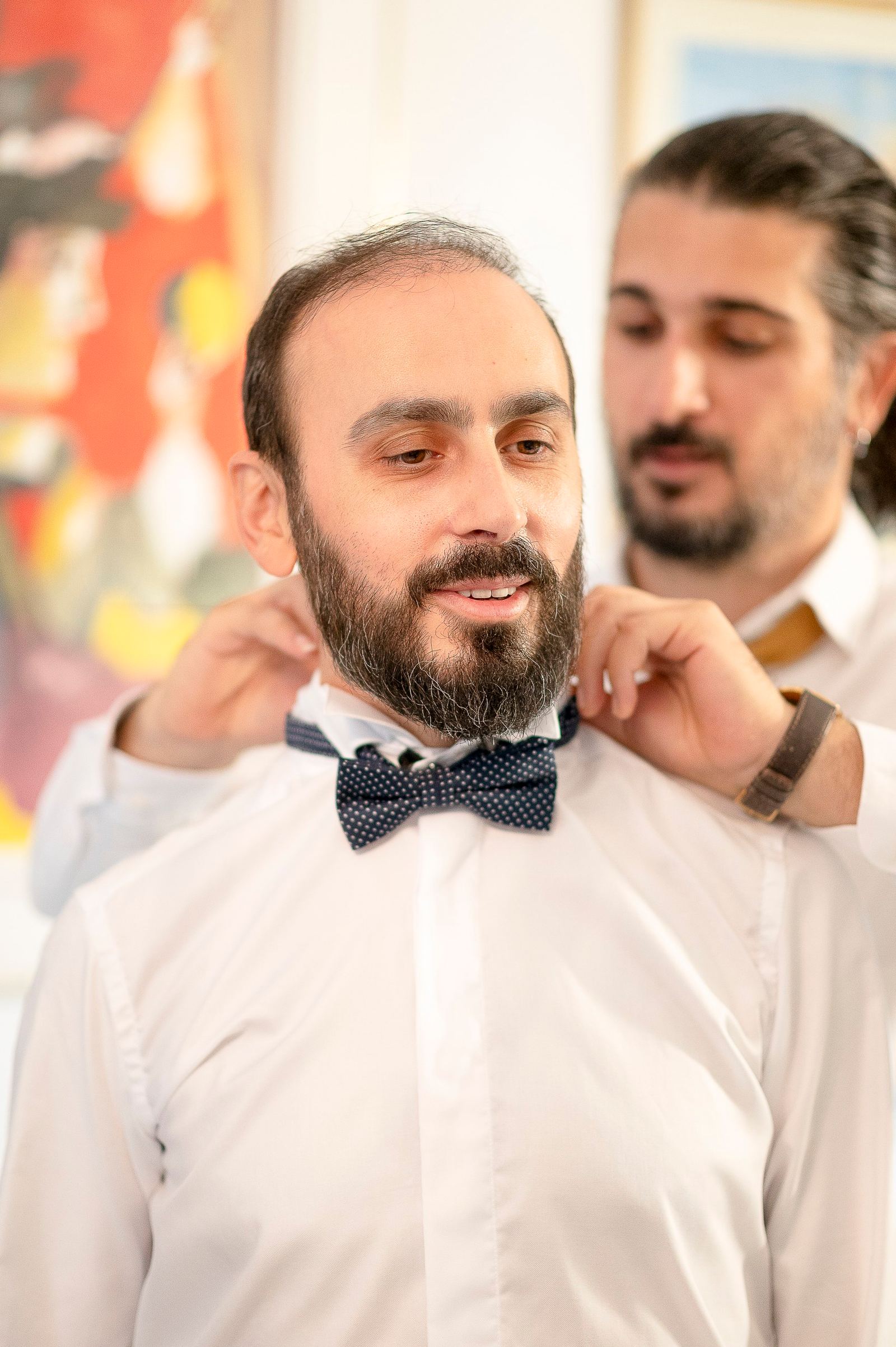 Ο γαμπρός χαμογελά καθώς ένας άντρας πίσω του φτιάχνει το πουά παπιγιόν του, με πολύχρωμο πίνακα πίσω.