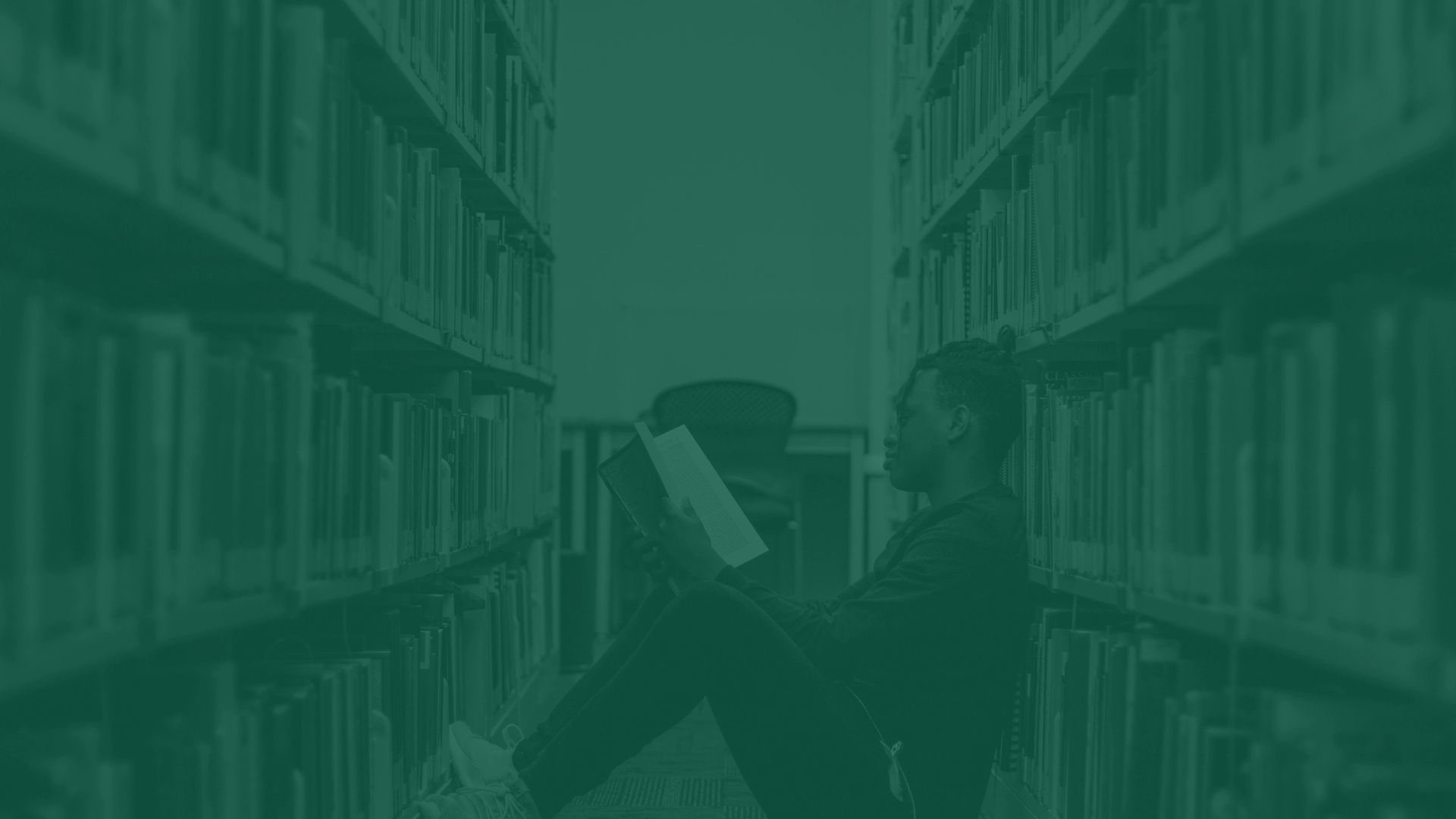 A person sitting on the floor reading in a library aisle.