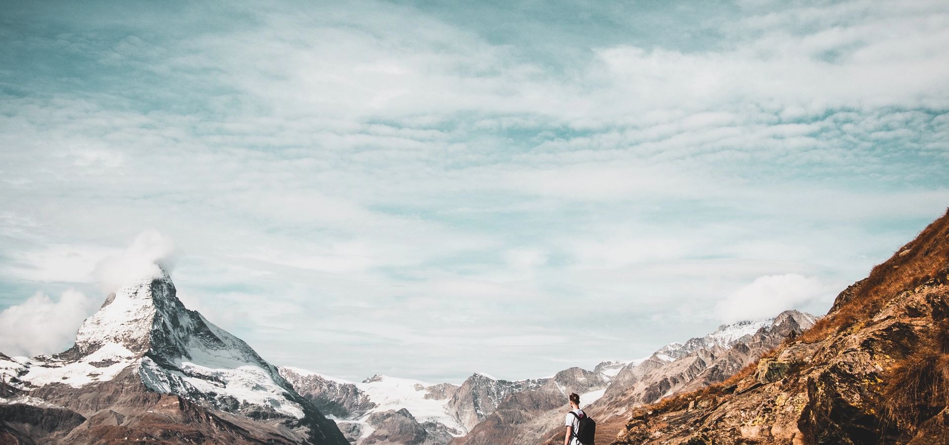 A hiker on a mountain trail looking out toward the Matterhorn under sweeping clouds — bold, values-driven ambition.