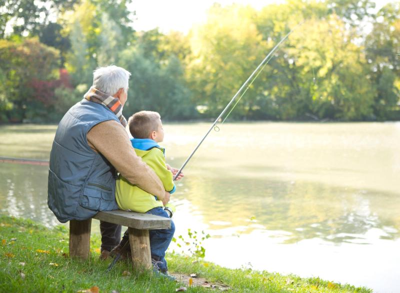 An older man and a young boy fishing together from a bench beside a lake