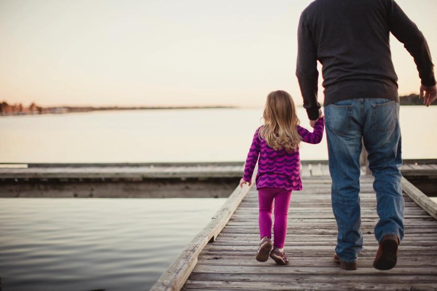A father and his young daughter walking hand in hand along a lakeside dock at sunset