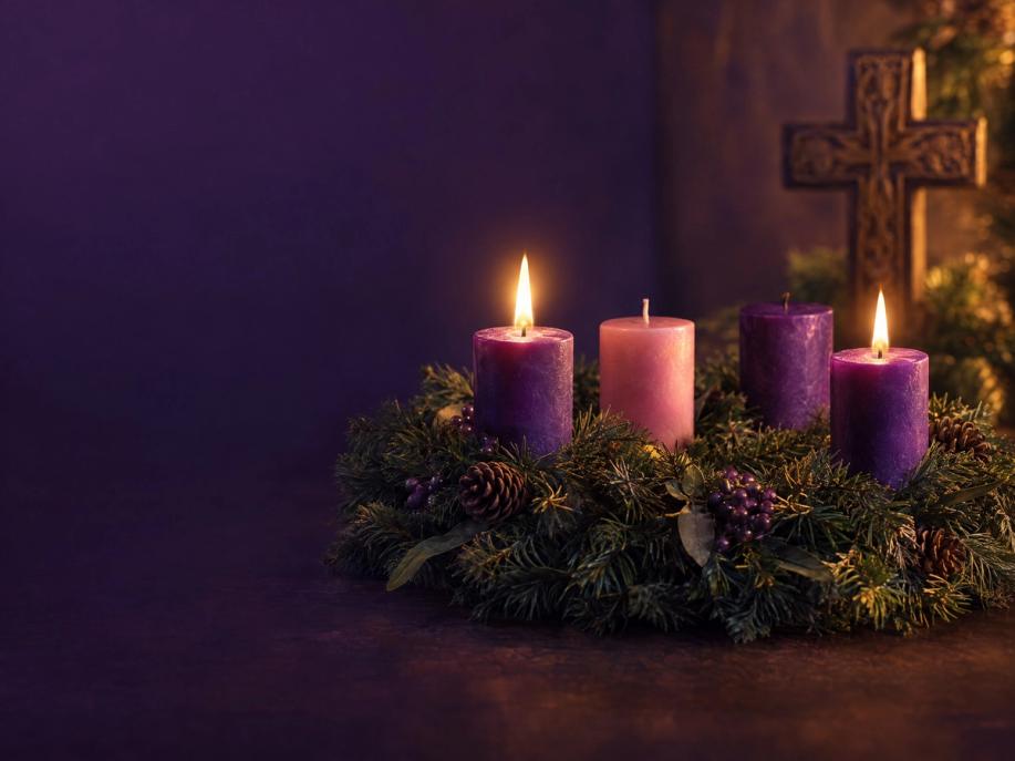 An evergreen Advent wreath with three violet candles and one rose candle, two lit, beside a carved wooden cross.