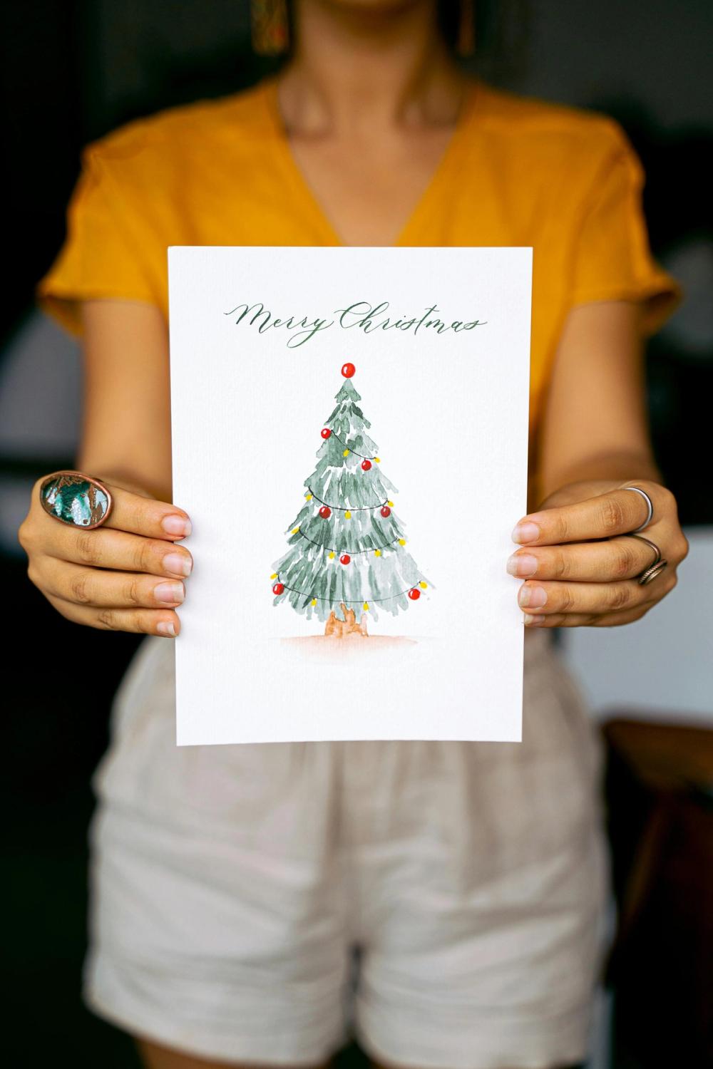 A woman holds out a white card with a hand drawn Christmas Tree illustration and the message "Merry Christmas"