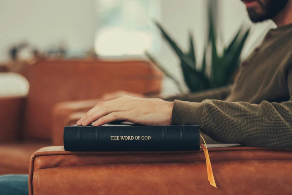 A black leather Bible resting on the arm of a sofa with a hand placed atop it