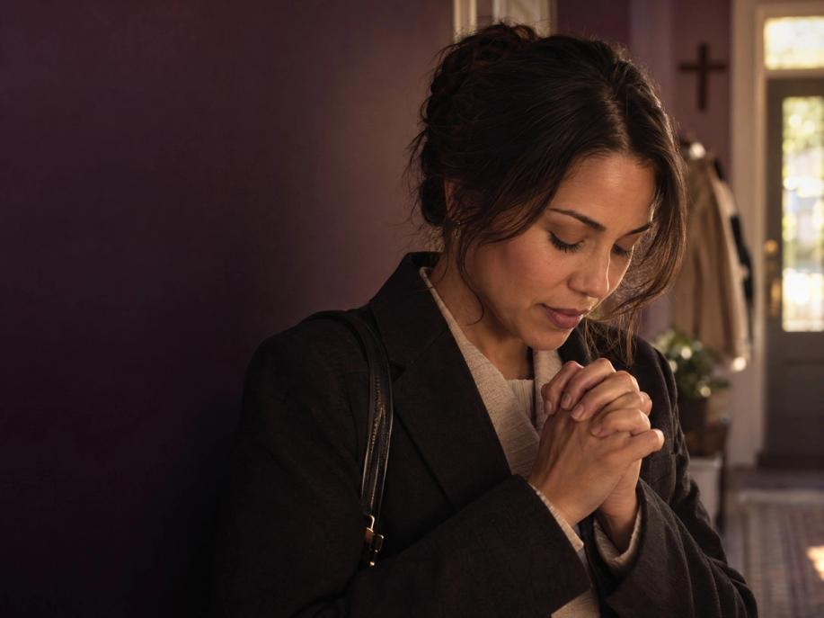 An adult in everyday clothing pauses at home with bowed head and clasped hands in prayer.