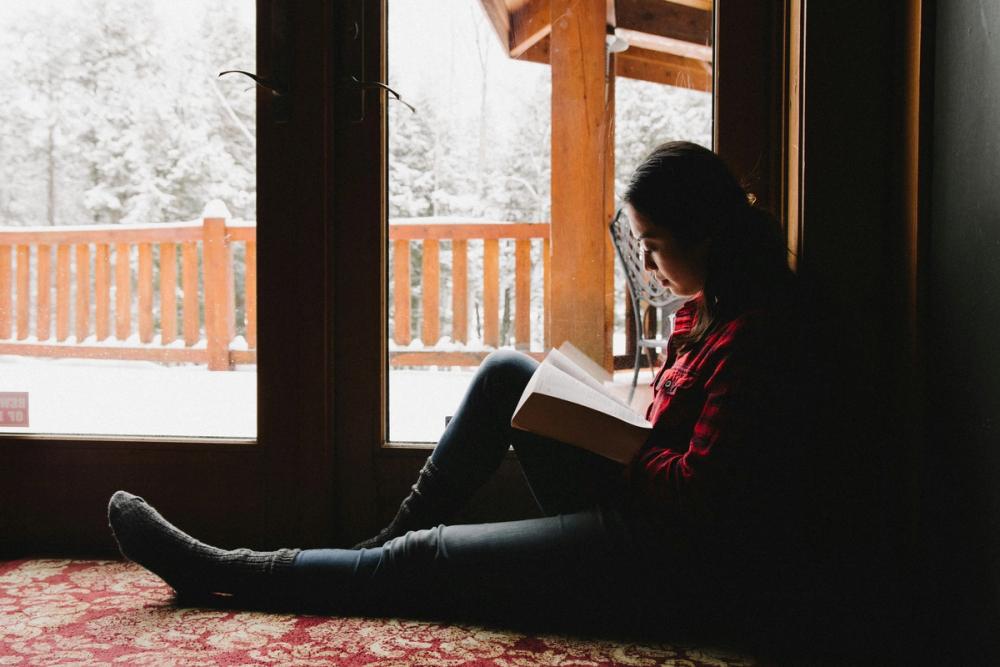 woman reading a Bible in a log cabin in the winter, it is snowing outside but cosy inside