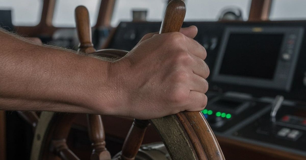 A close-up, photorealistic image of a seafarer's hands gripping a worn steering wheel, conveying the pressure of the job.