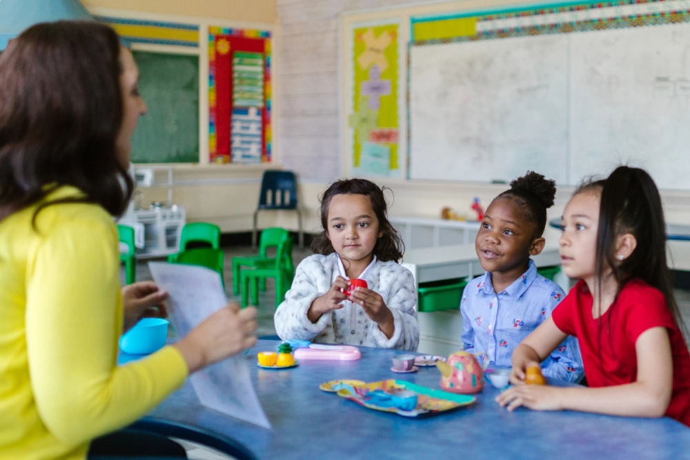 A teacher readers to children in a classroom. Three girls of varying race and ethnicities