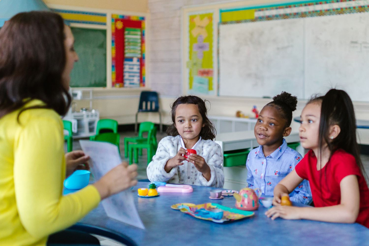 A teacher readers to children in a classroom. Three girls of varying race and ethnicities