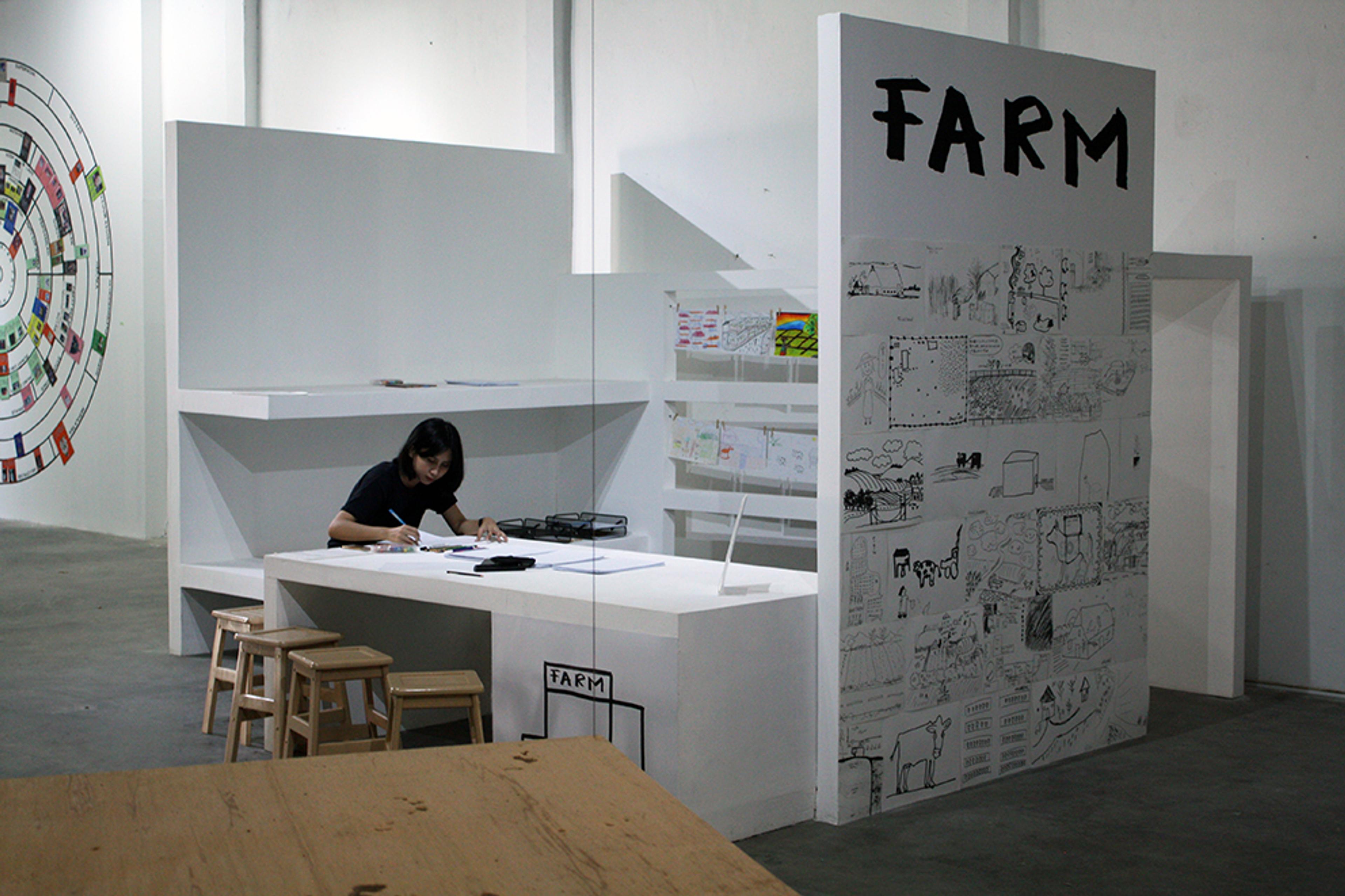 A room-size booth with three walls and a table inside, the outdoor walls are covered with handdrawings, and the word FARM is written in large letters. Inside the booth is a person sitting at the table, and drawing.