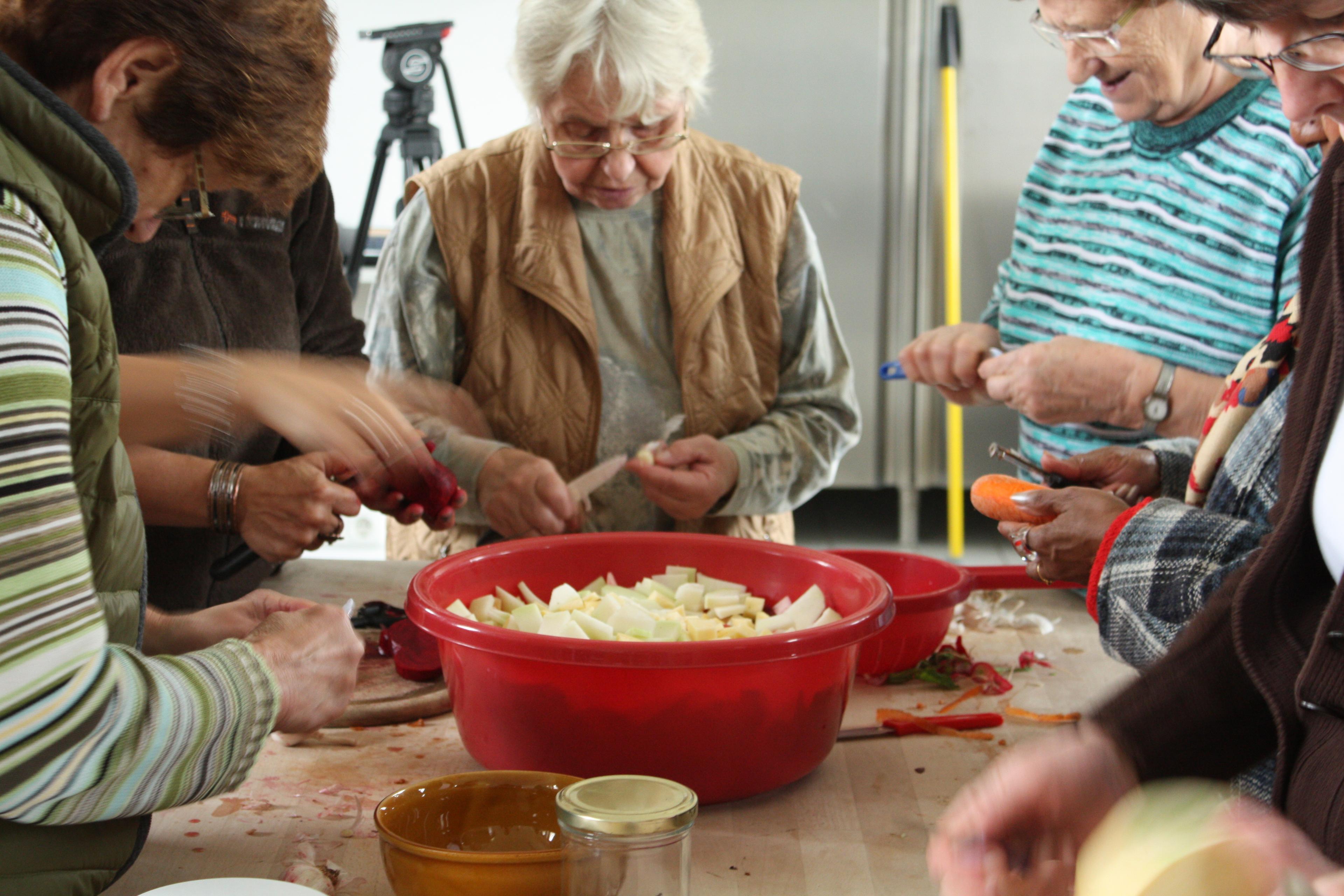 A group of women are places around a big red bowl on a wooden table. They all peel and chop vegetables.