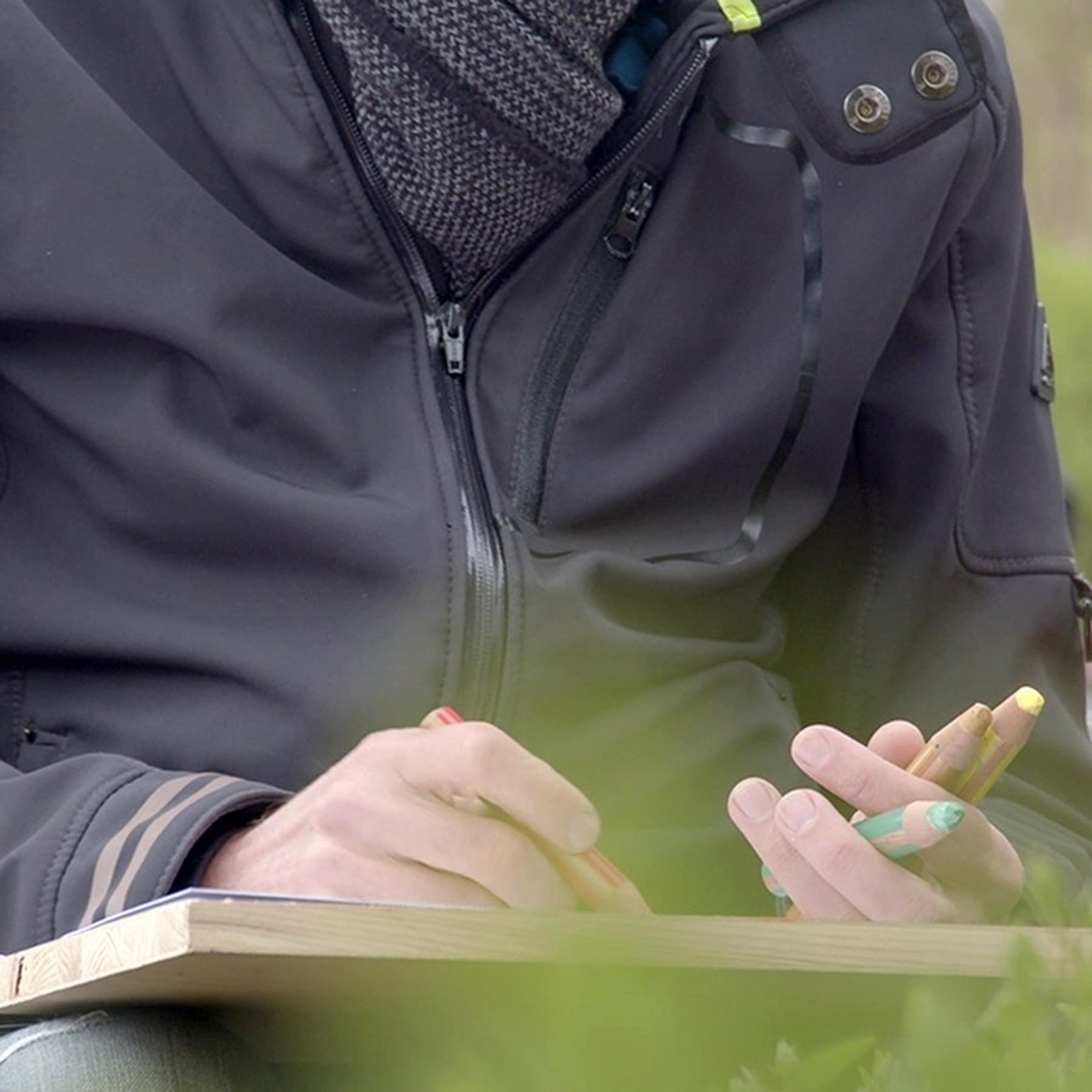 Cropped image of a person sat drawing. Their left hand holds three colour crayons, their right hand draws with a red crayon on paper on a board resting on their lap.
