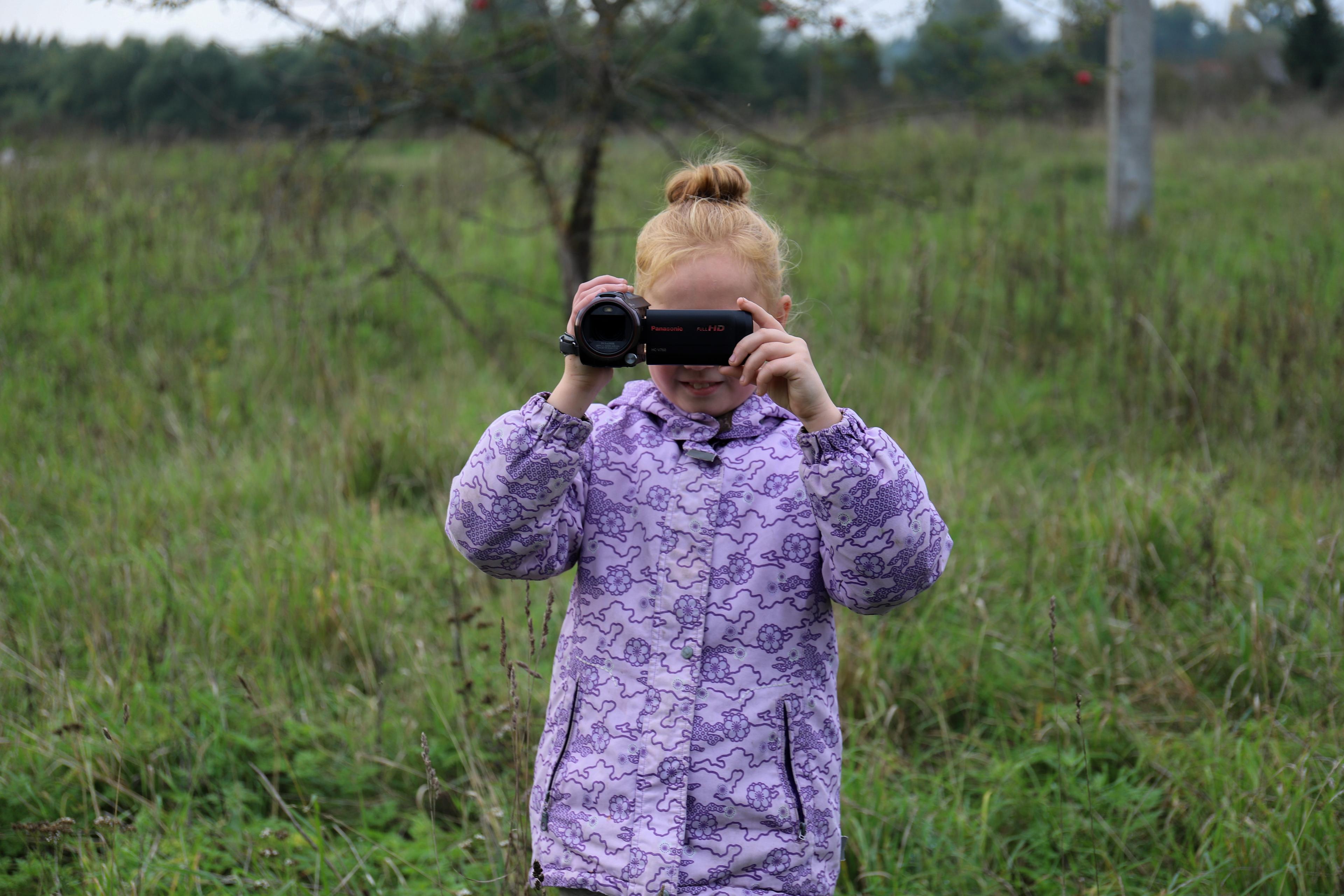A girl holding a video camera in front of a field. The camera obscures her face.