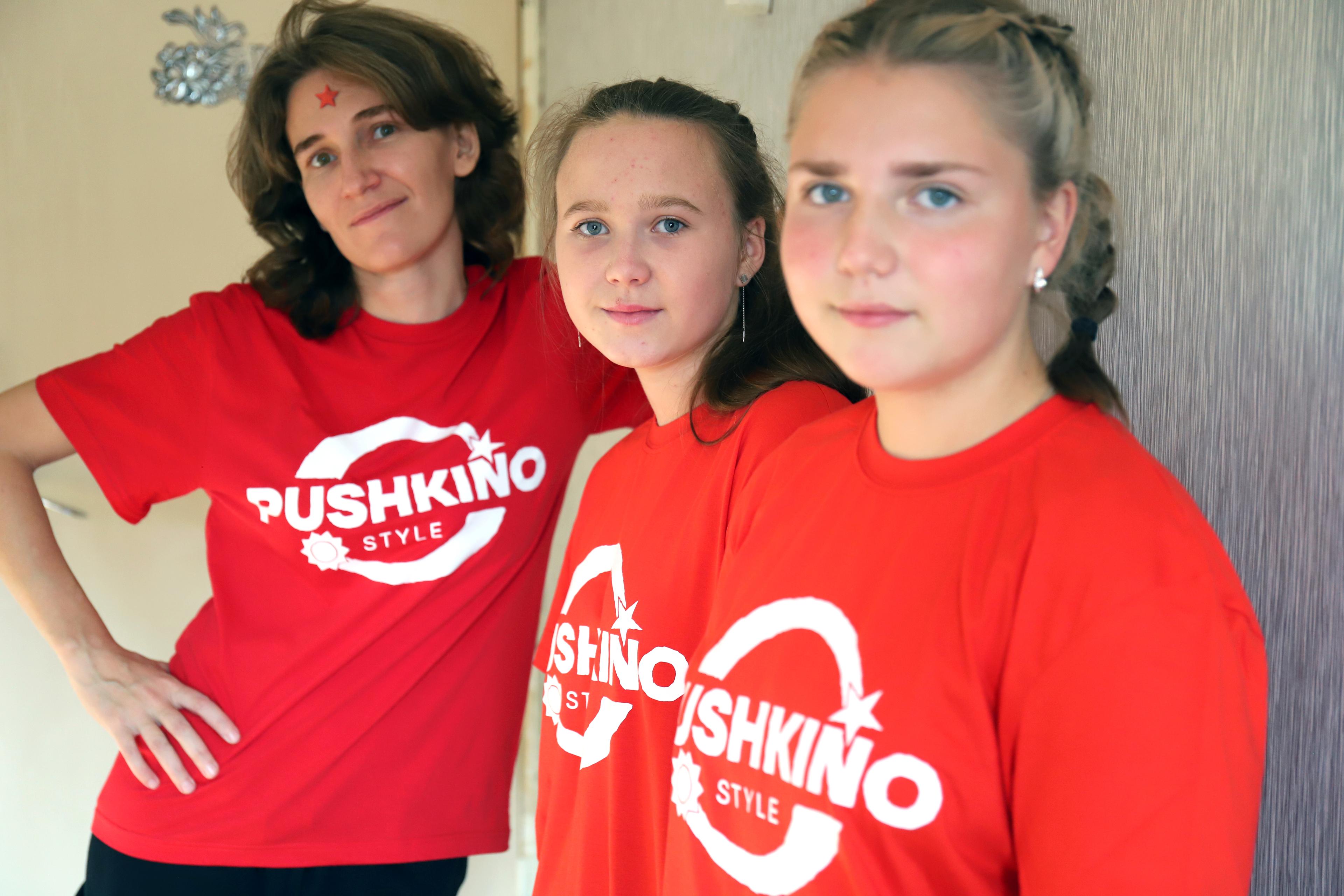 A women and two girls are leaning against a wall and are looking straight into the camera. Thea are wearing bright red T-shirts with Pushkino Style Logo printed in white on the front.